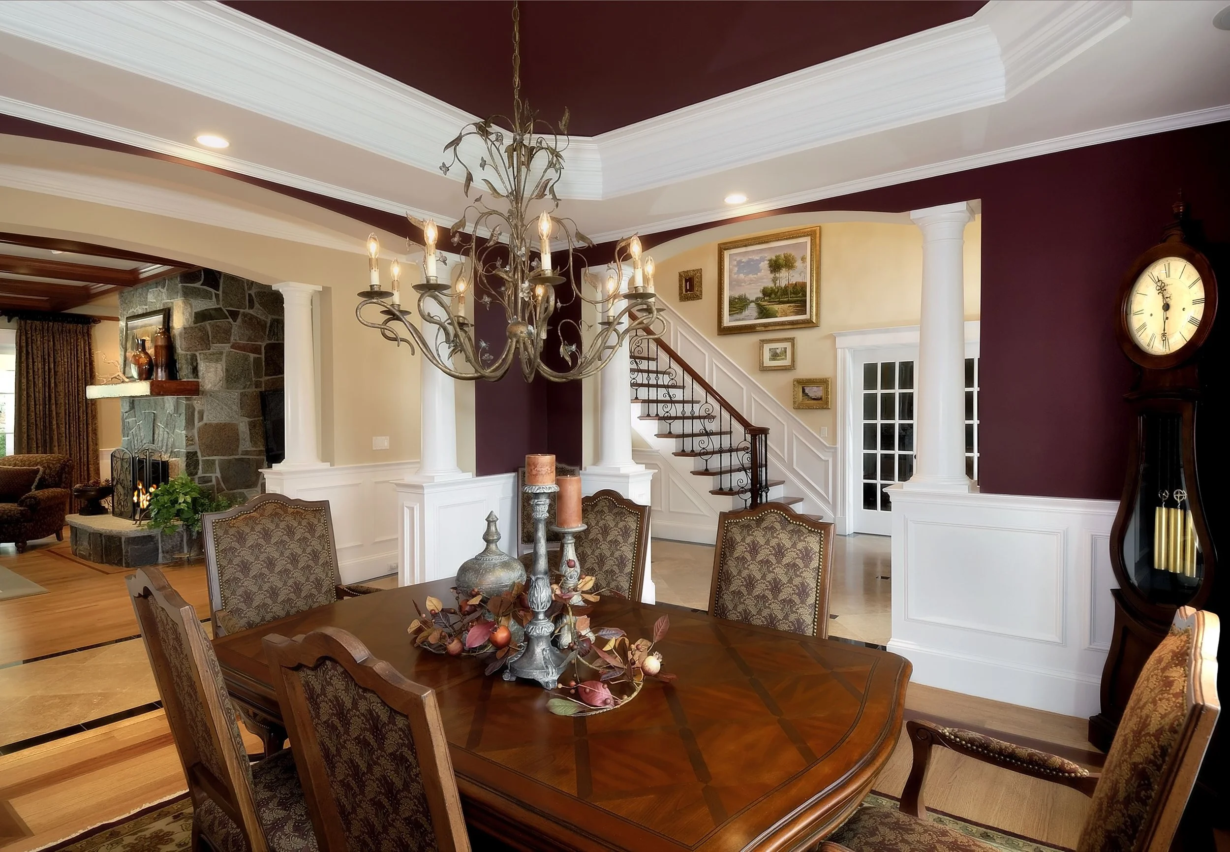 Elegant dining room with a wooden table and chairs, a chandelier, and decorative items on the table. The room has white trim, columns, and framed artwork on the walls. A staircase with a wooden handrail and wrought iron balusters leads upstairs, and 