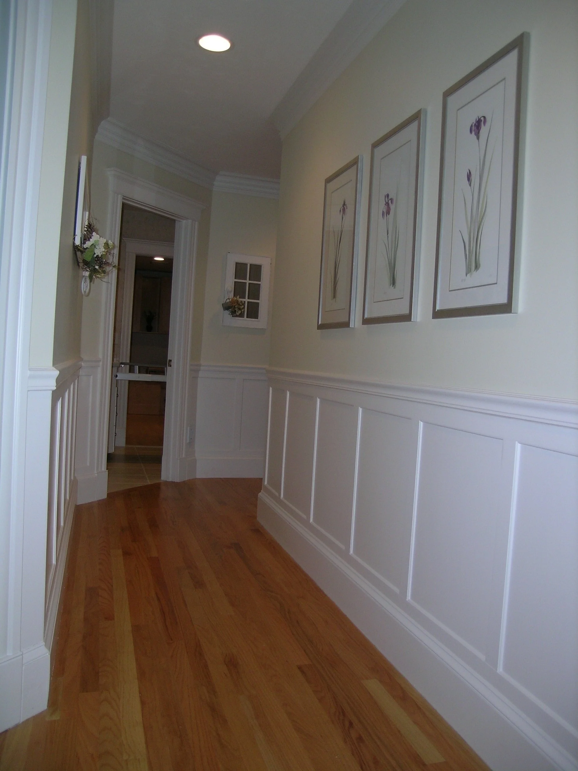 A hallway with hardwood floors, paneled wainscoting on the walls, three framed botanical prints, a small wall-mounted flower arrangement, and an open door leading into a room with a kitchen visible in the background.
