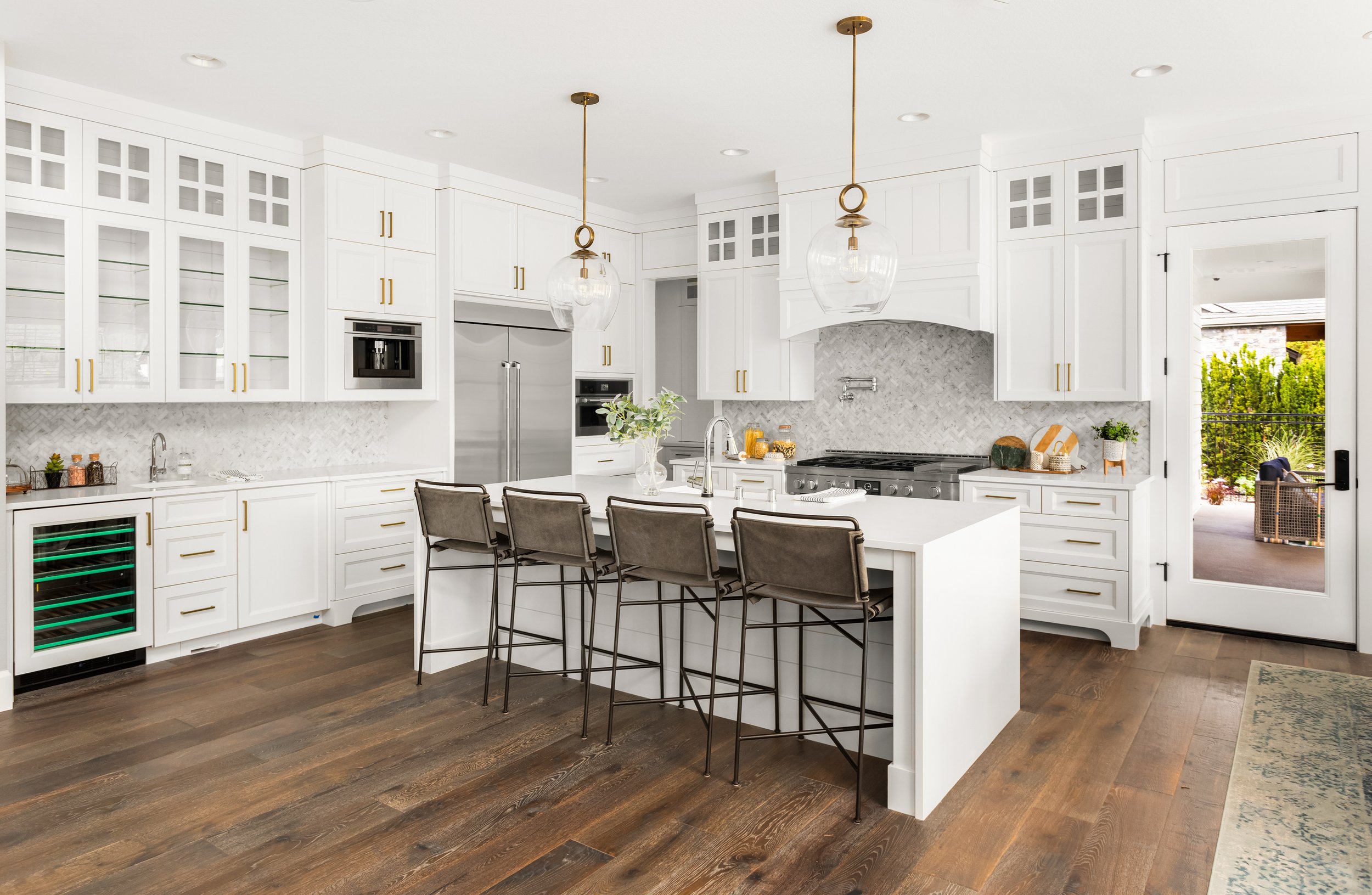 Modern white kitchen with dark wood flooring, kitchen island with four chairs, stainless steel appliances, pendant lights, and a glass door leading outside.