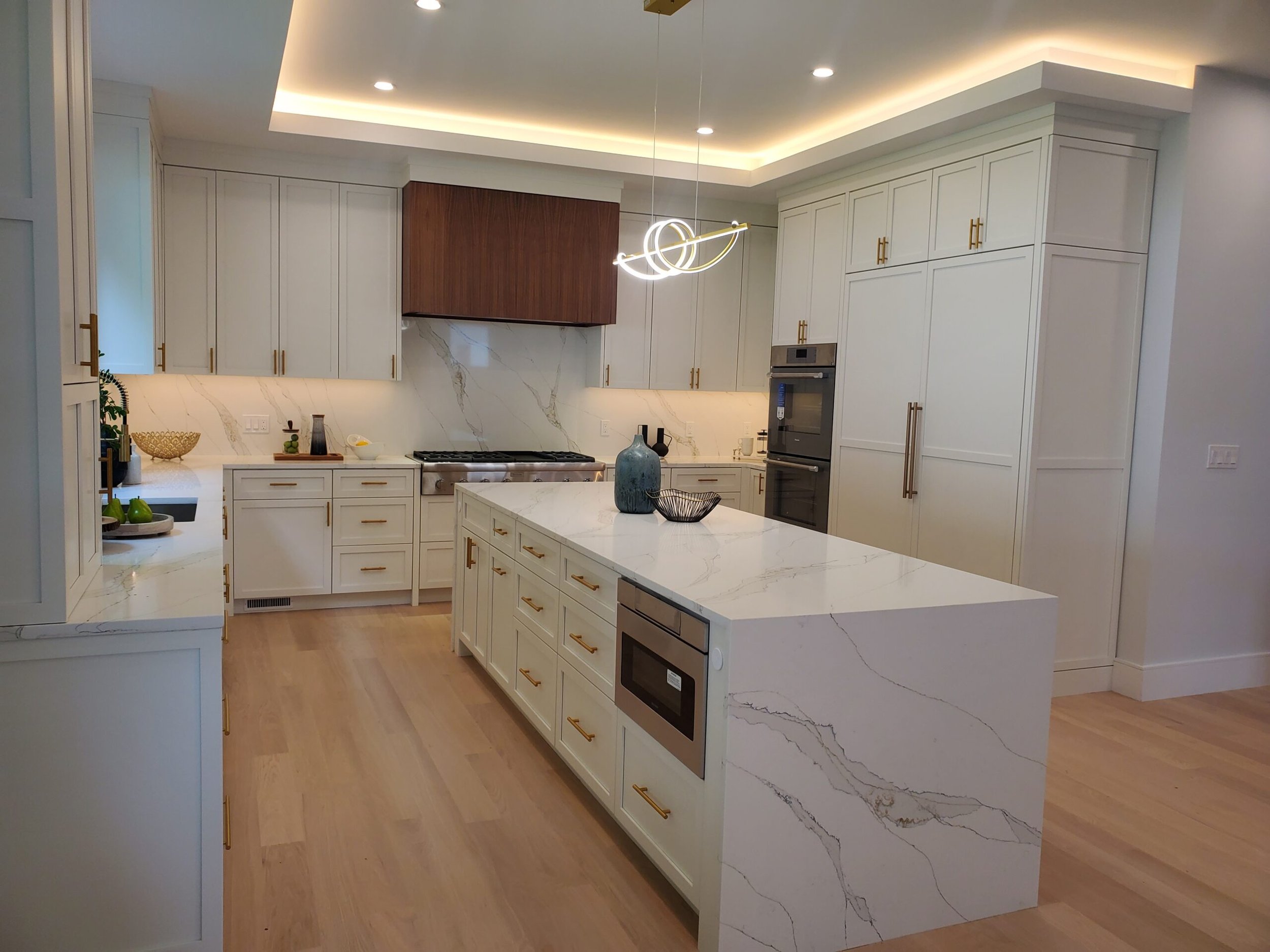 Modern kitchen with white cabinets, marble countertops, and wooden accents. A central island features a built-in microwave and decorative vases. Pendant lighting and under-cabinet lighting illuminate the space.