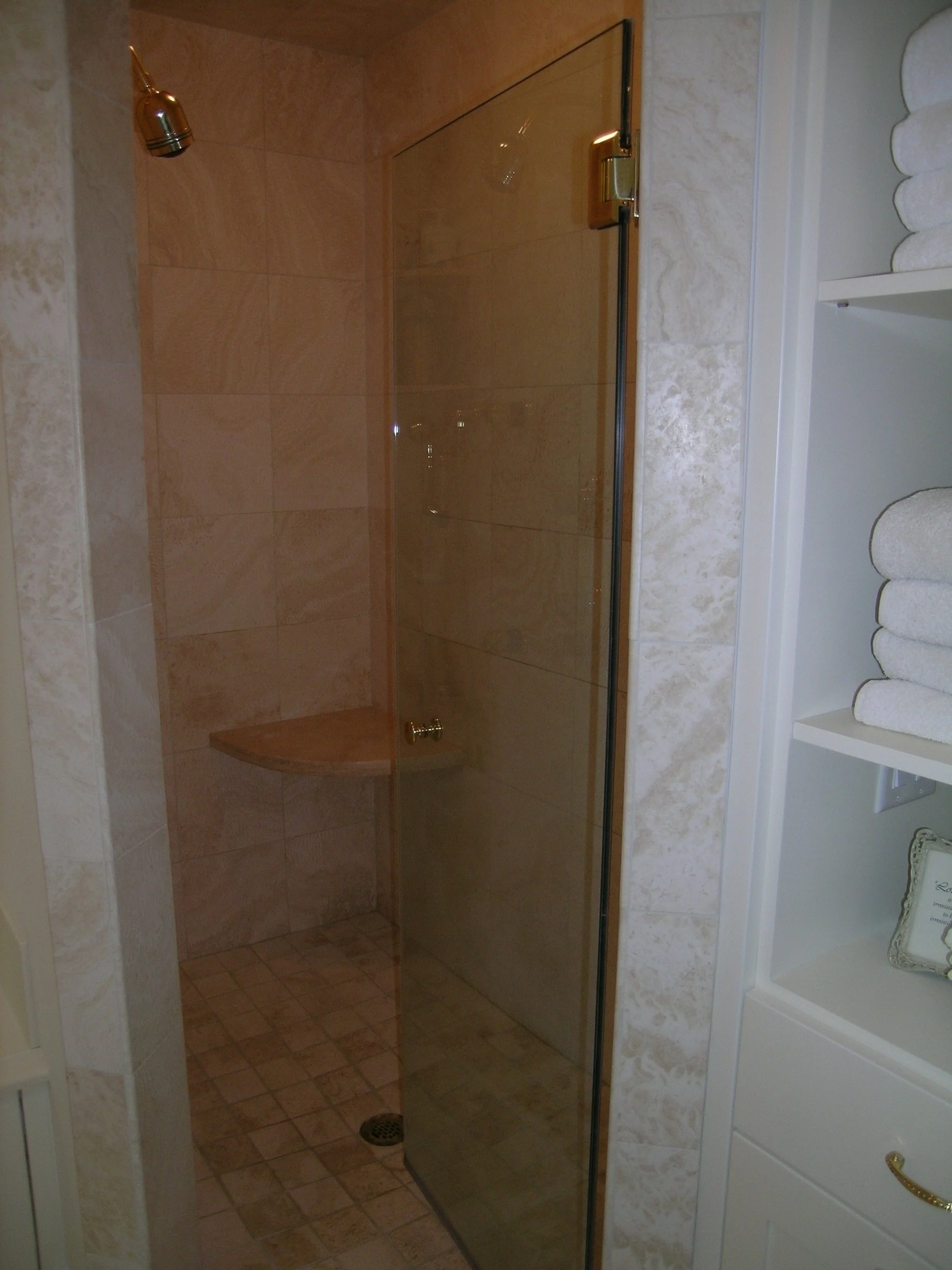 A small shower area with beige tile walls, a brass showerhead, a built-in corner shelf, and a glass door, next to a cabinet with neatly folded white towels.