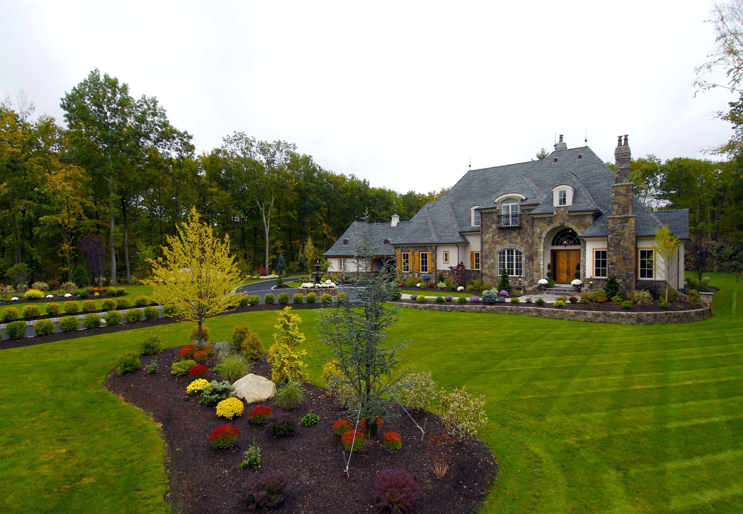 Large house with stone and white exterior, steep dark roof, and multiple chimneys, surrounded by well-maintained green lawn, colorful garden, and trees in the background.