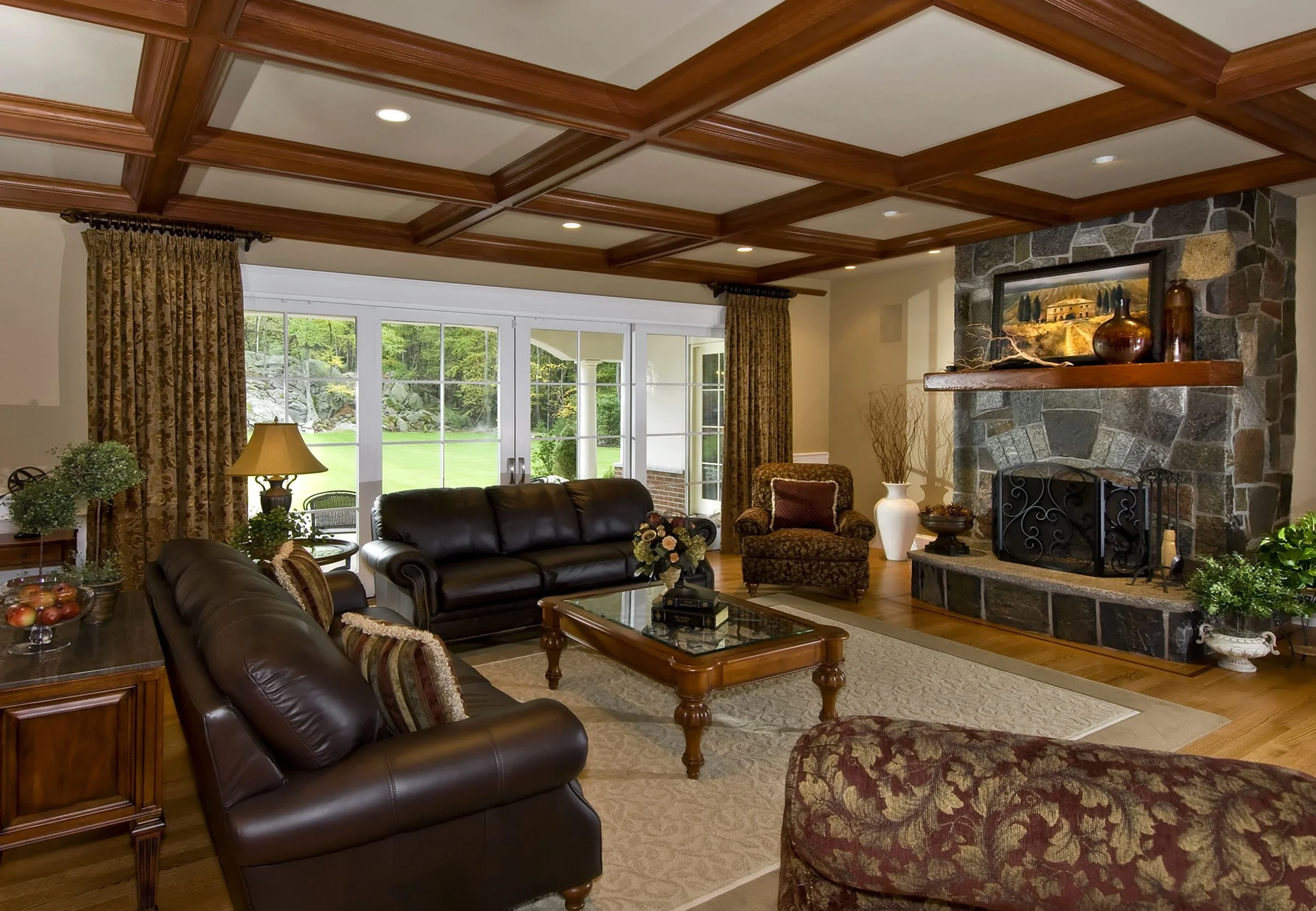 Living room with wood-paneled coffered ceiling, stone fireplace with a wooden mantel, leather sofas, patterned armchair, large windows overlooking a green yard, and various decorative items like vases and a painting.