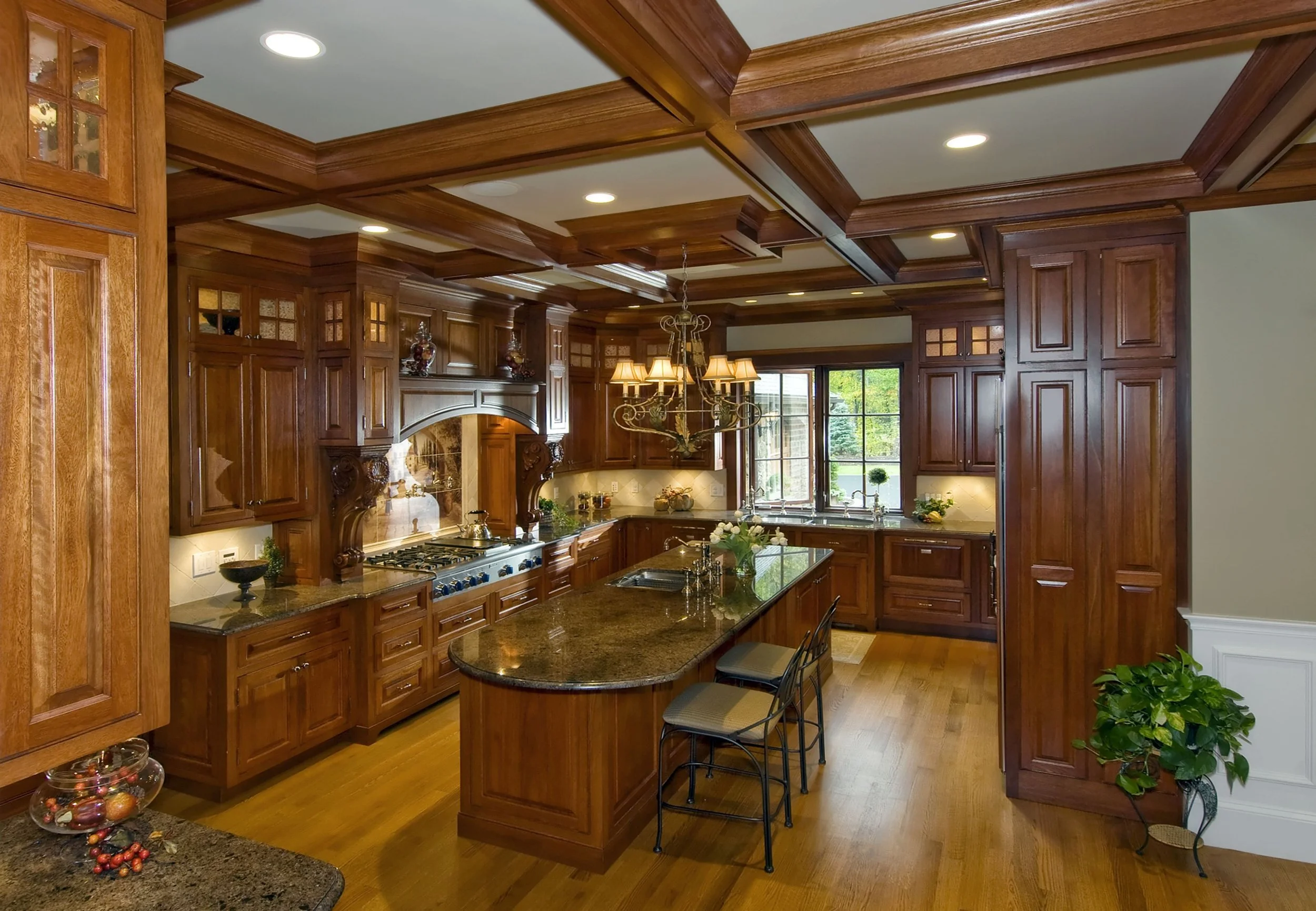Large kitchen with wooden cabinetry, granite countertops, and a central island with seating. A chandelier hangs above the island. A window above the sink shows greenery outside. The ceiling has detailed wooden molding, and the floor is hardwood. Smal