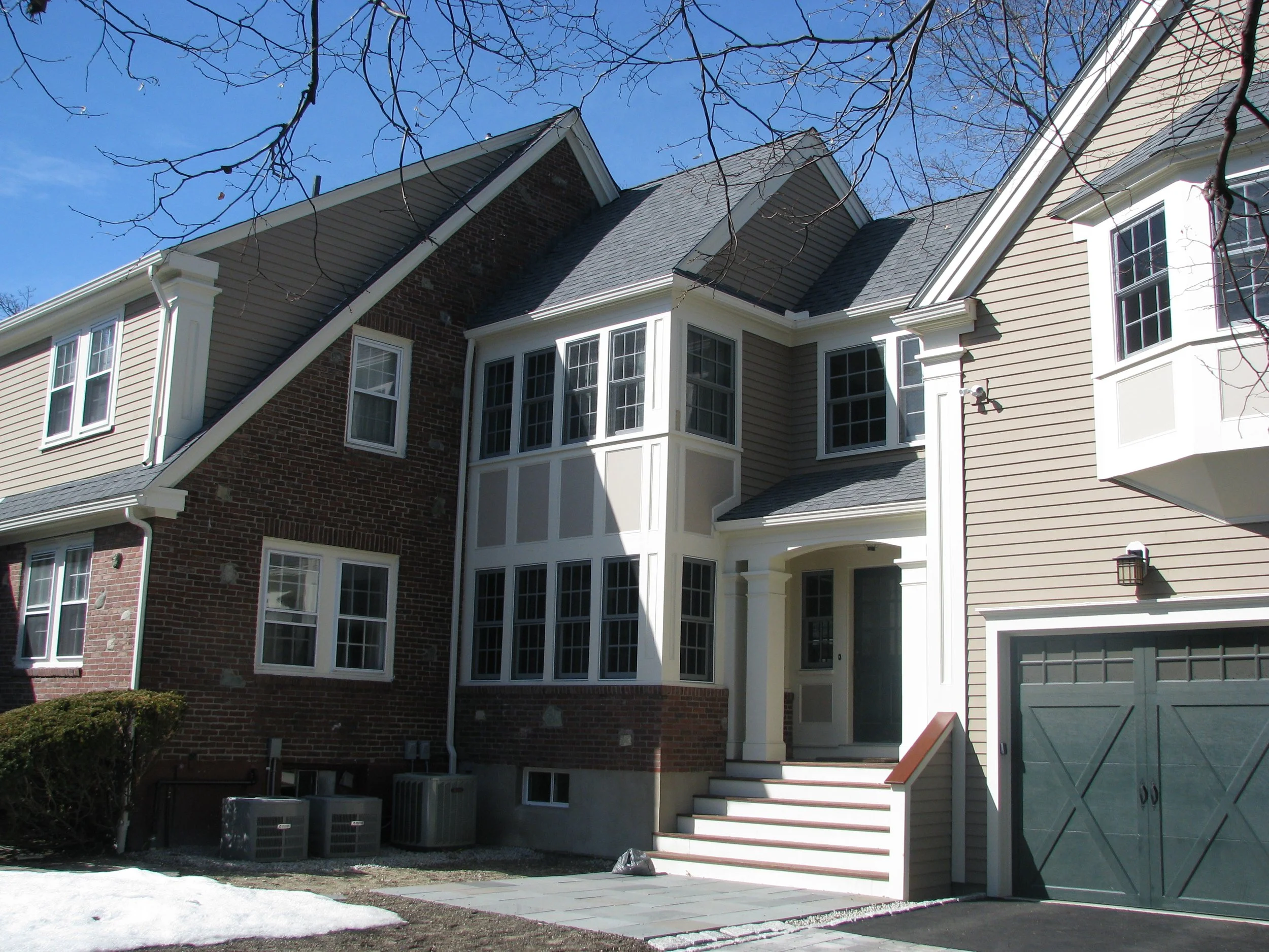 Modern suburban house with brick and siding exterior, multiple windows, a front porch with stairs, and an attached garage. Snow visible on the ground.