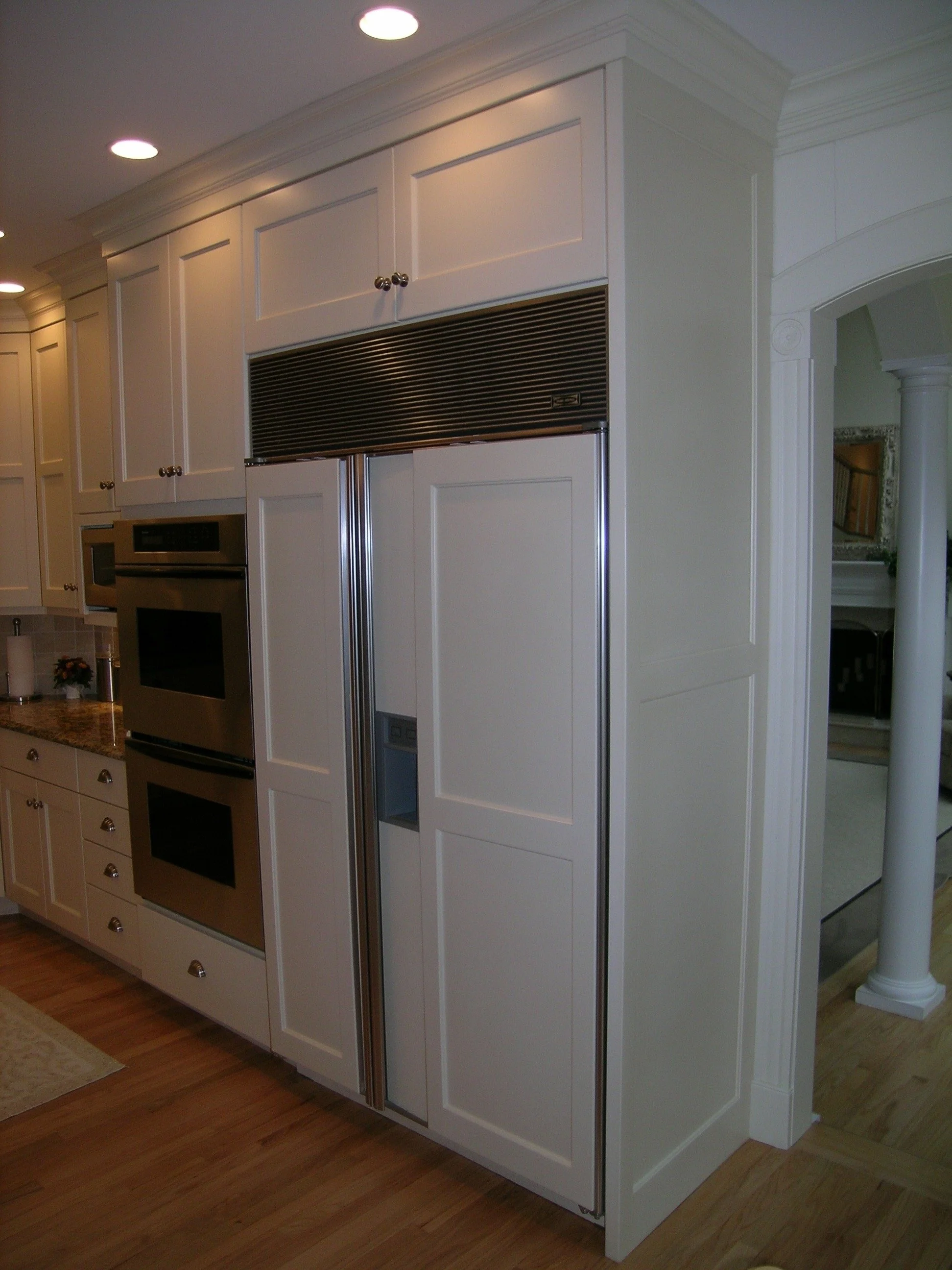 Modern kitchen with built-in oven and refrigerator, white cabinets, oak flooring, and recessed lighting.