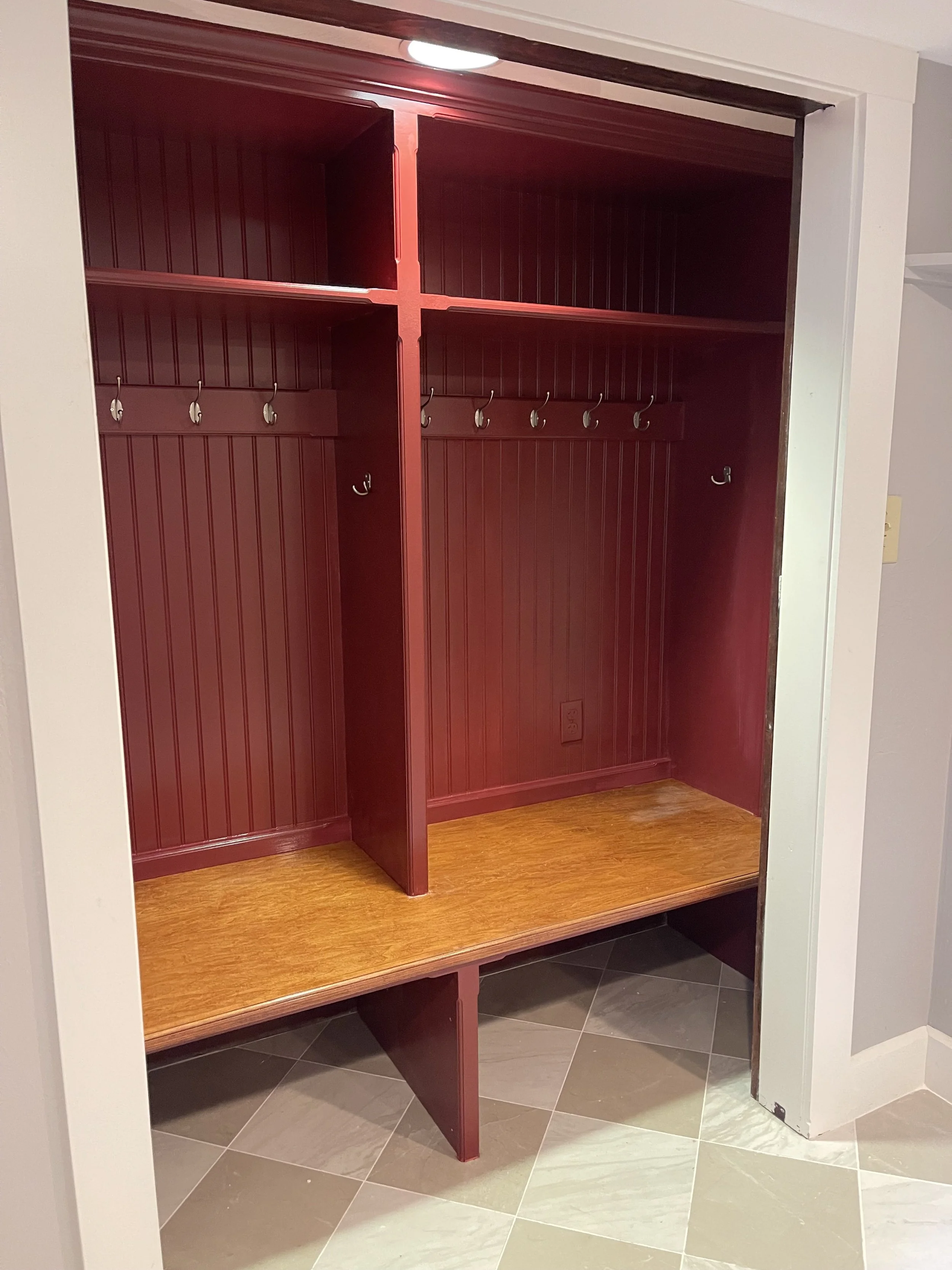 Empty red wooden closet with hooks on the back wall and a wooden bench at the bottom, situated in a room with tiled floor.