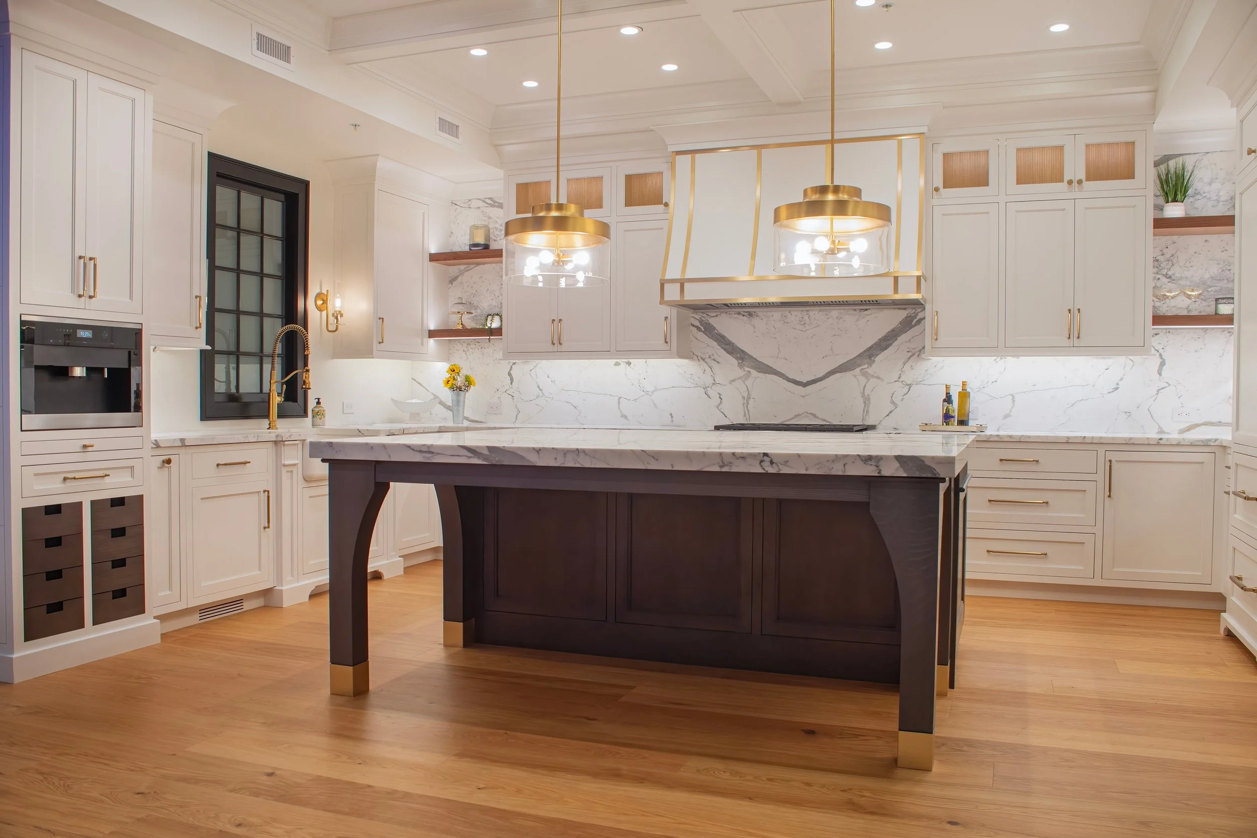 Modern kitchen with white cabinetry, marble backsplash and countertops, a black window, wooden shelves, a dark island with gold accents, and three gold pendant lights hanging above the island.