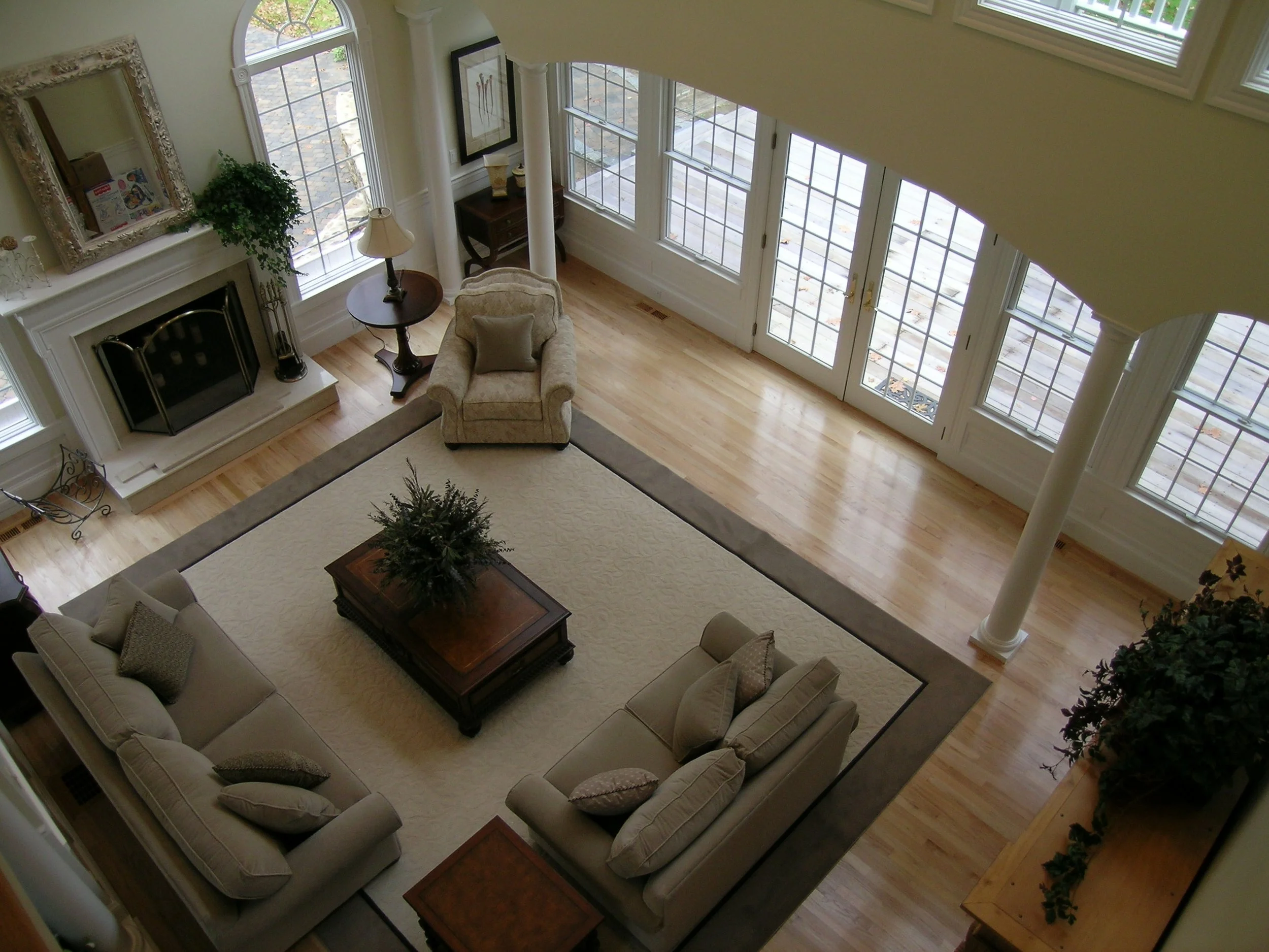 A living room with large windows, hardwood floors, a fireplace, a beige sofa with cushions, an armchair, a coffee table with a plant on it, and a side table with a lamp, seen from above.