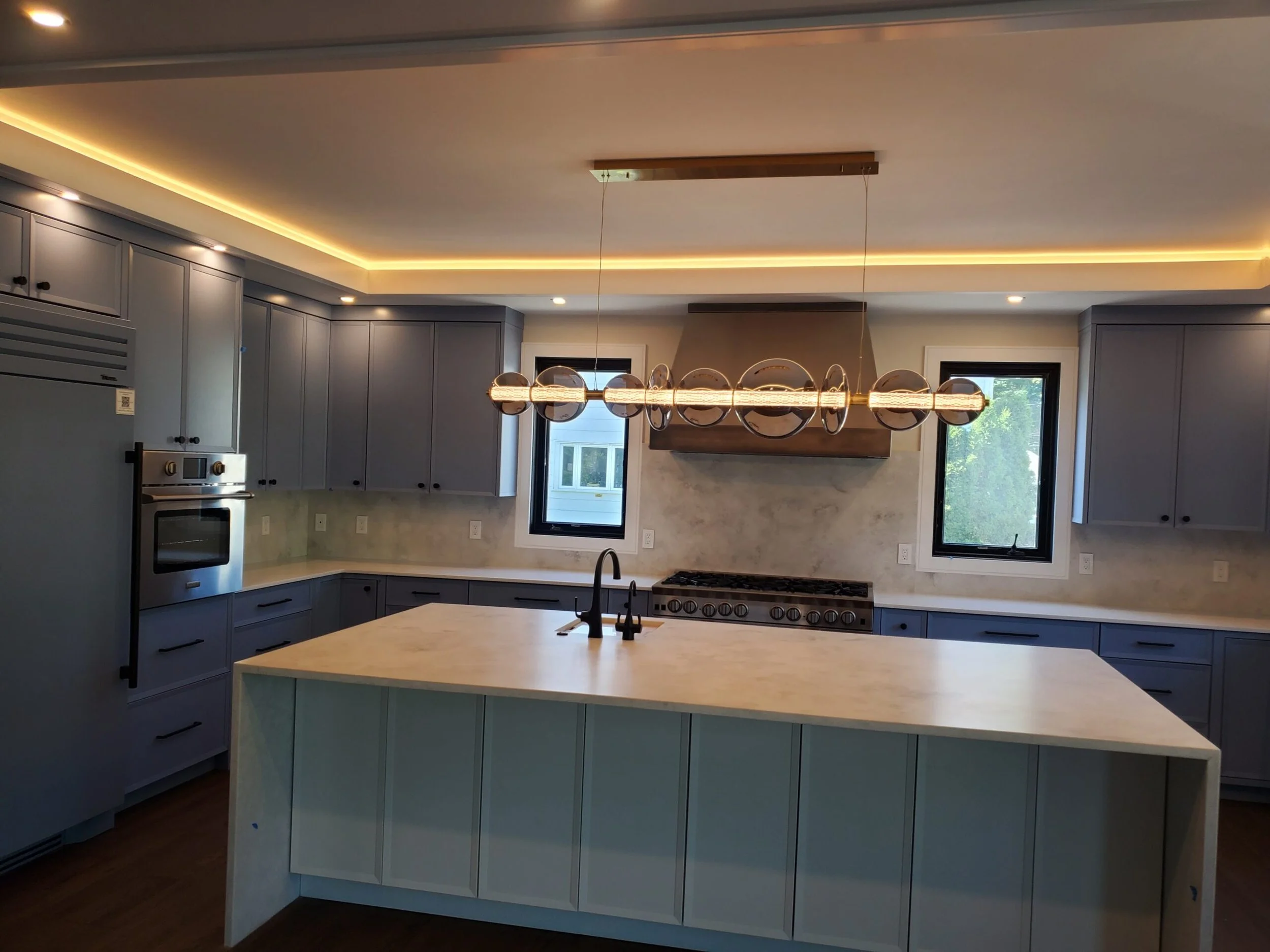 Modern kitchen with gray cabinets, marble backsplash, large island with a sink, and unique chandelier lighting fixture.