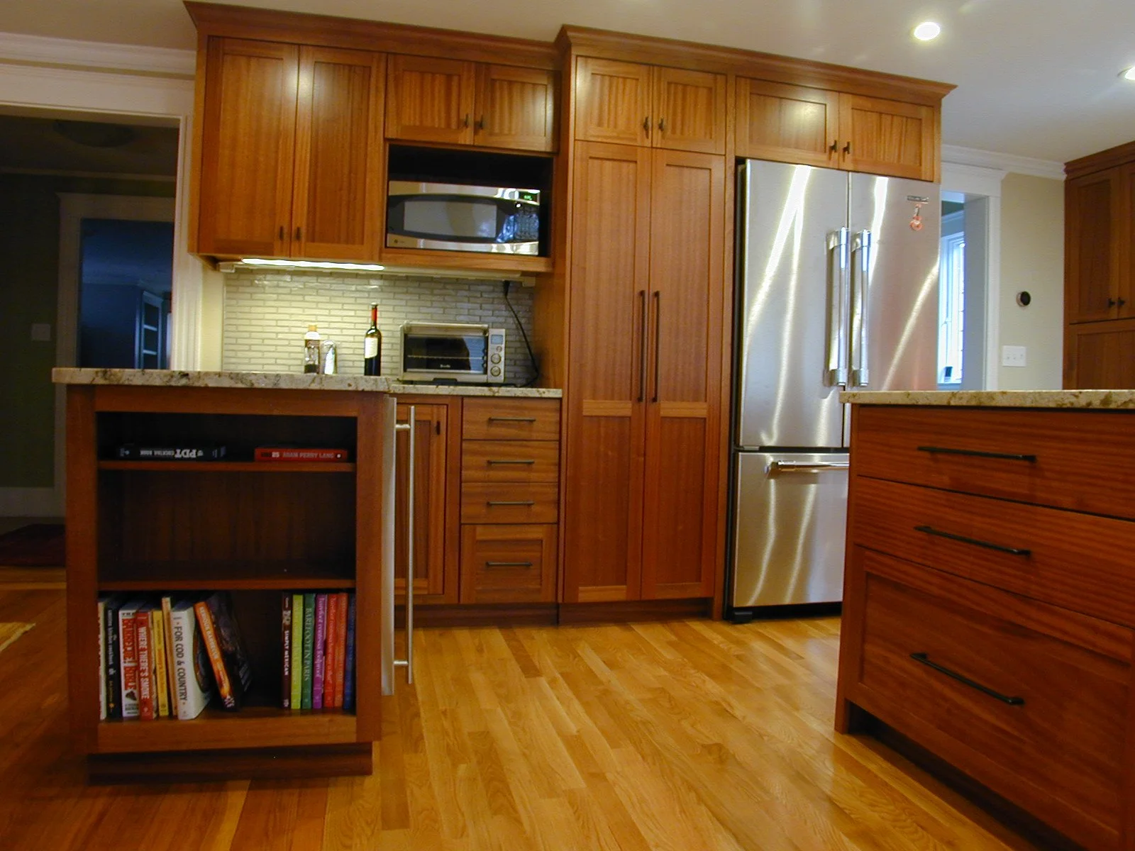 Kitchen with wooden cabinets, stainless steel refrigerator, microwave, toaster oven, bookshelves, and hardwood flooring.