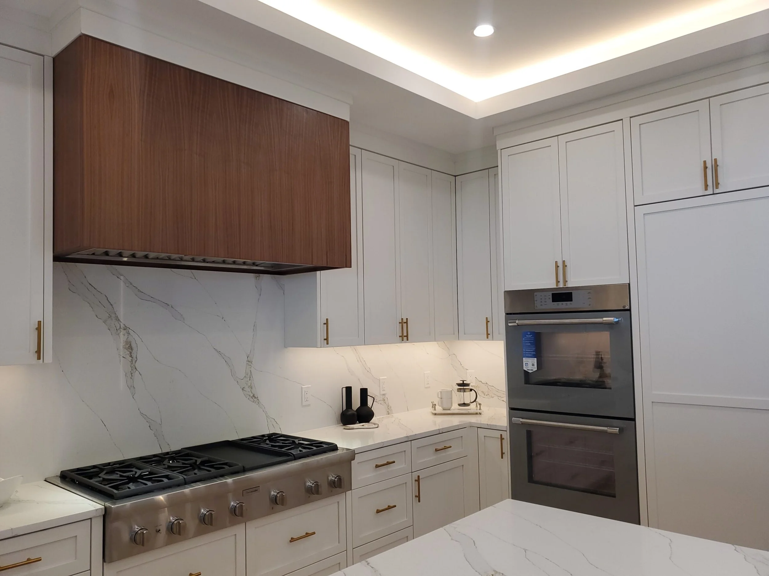Modern kitchen with white cabinets, marble backsplash, and marble countertops. Stainless steel double wall oven, gas stove with five burners, and decorative black vases on the counter. Wooden range hood above the stove, and ceiling with recessed ligh
