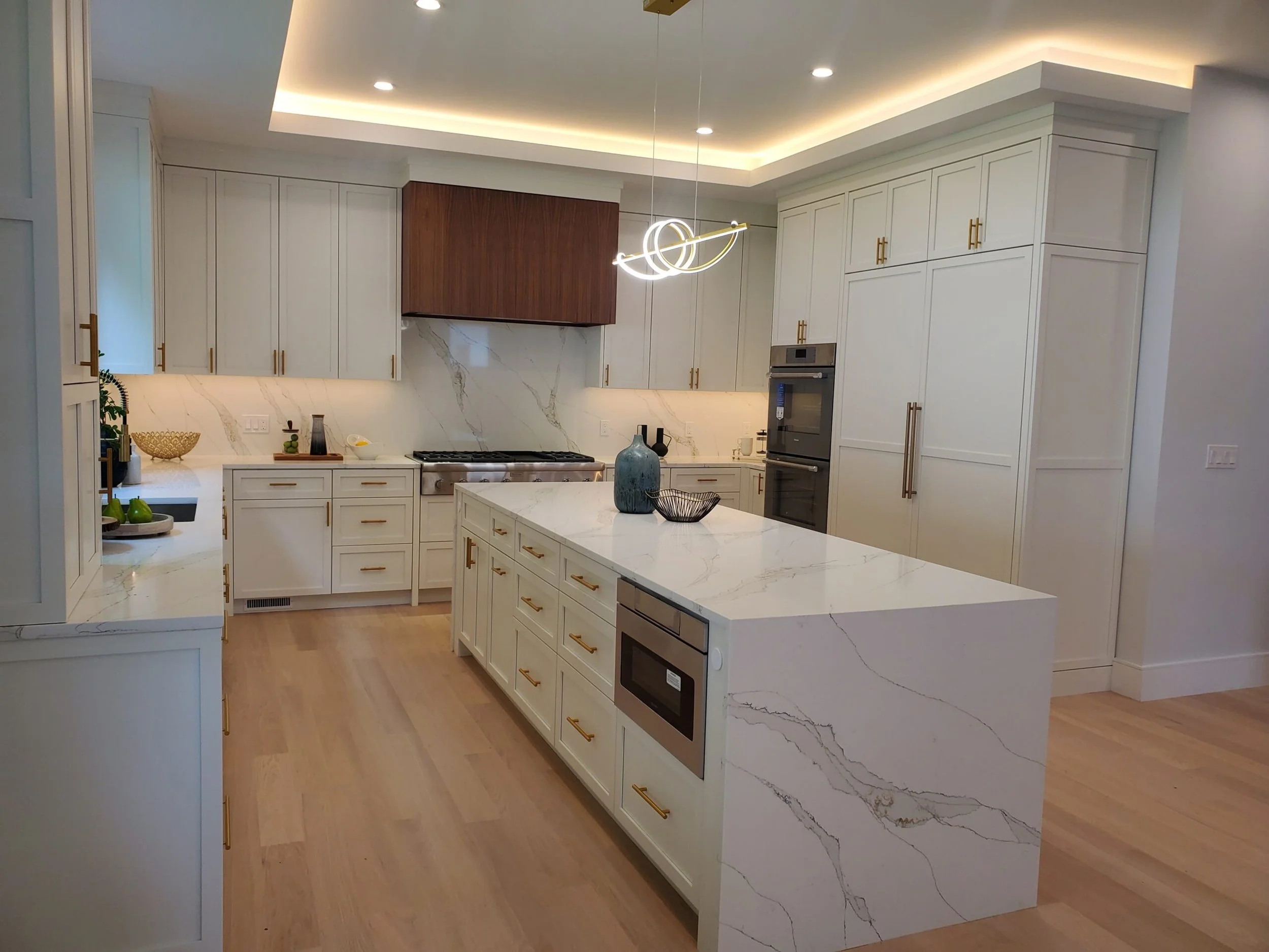 Modern kitchen with white cabinets, gold handles, marble countertops, a wooden range hood, and integrated appliances. A marble island with decorative vases and bowls. Hardwood floors and recessed ceiling lighting.