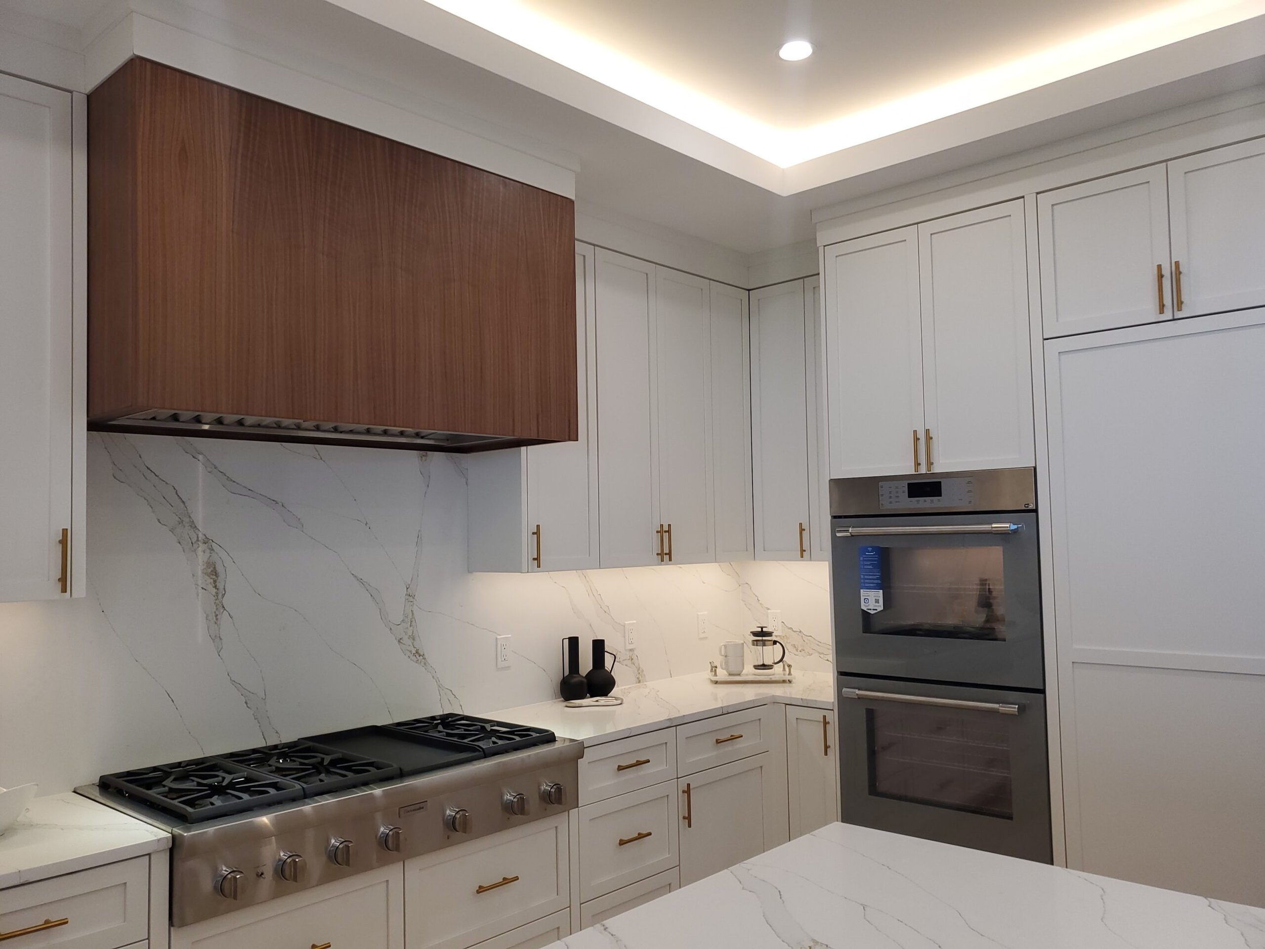 Modern kitchen with white cabinets, gold handles, marble backsplash, stainless steel oven, and hood. A small marble-topped island in foreground.