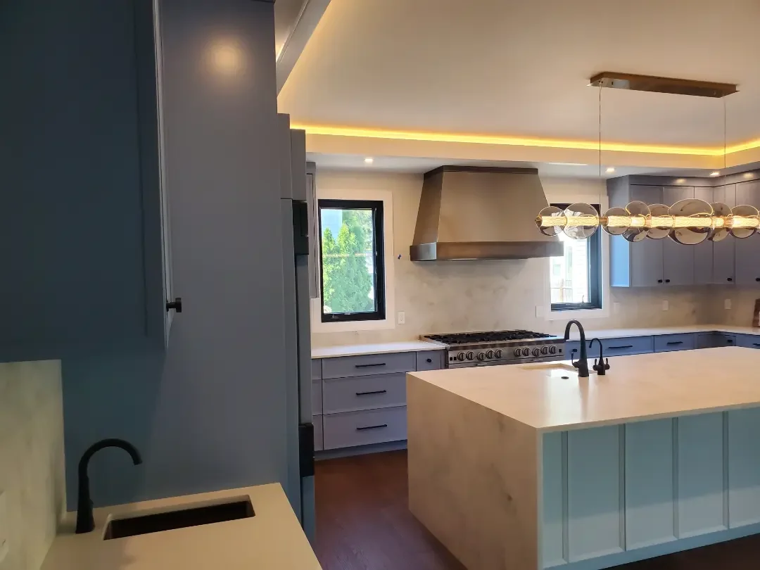 Modern kitchen with light gray cabinetry, a large island with a light-colored countertop, a black kitchen sink and faucet, stainless steel range hood, two small windows, a contemporary chandelier, and hardwood flooring.