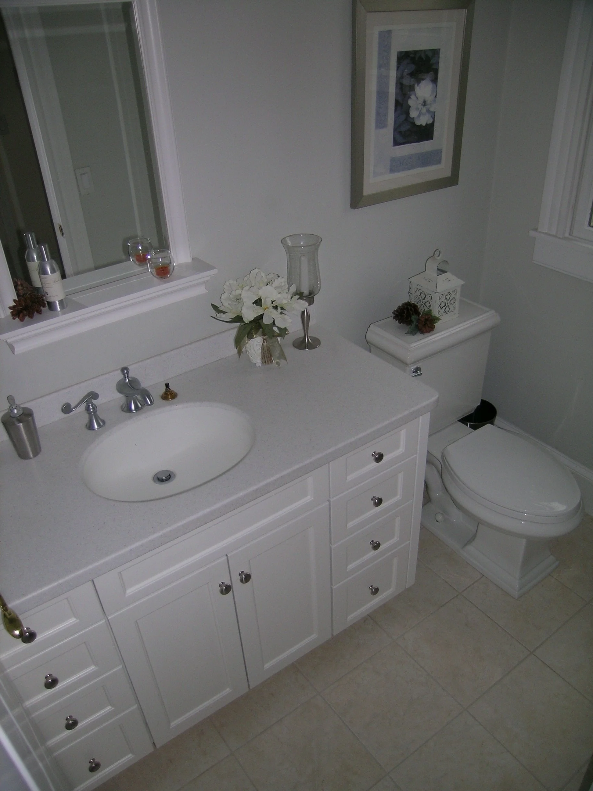 A bathroom with a white vanity, a vessel sink, a mirror, a framed picture of a white flower, and decorative items including a vase with white flowers, a tall glass candle holder, and a small lantern on the toilet tank. The toilet is positioned next t