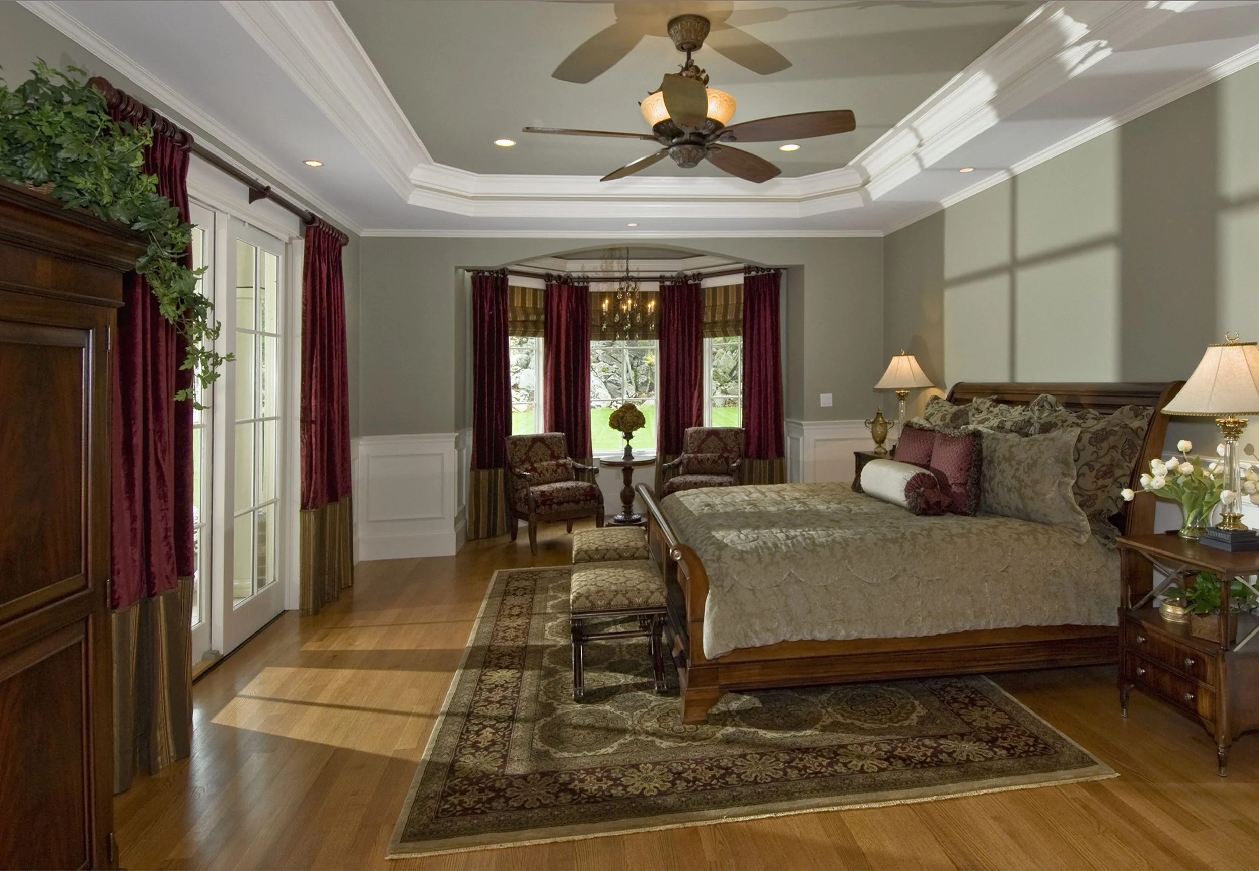 Spacious bedroom with cream-green walls, large windows with red and gold curtains, wooden furniture including a bed with patterned bedding, nightstands with lamps, a seating area with two chairs and a table, a wooden cabinet, and a ceiling fan.