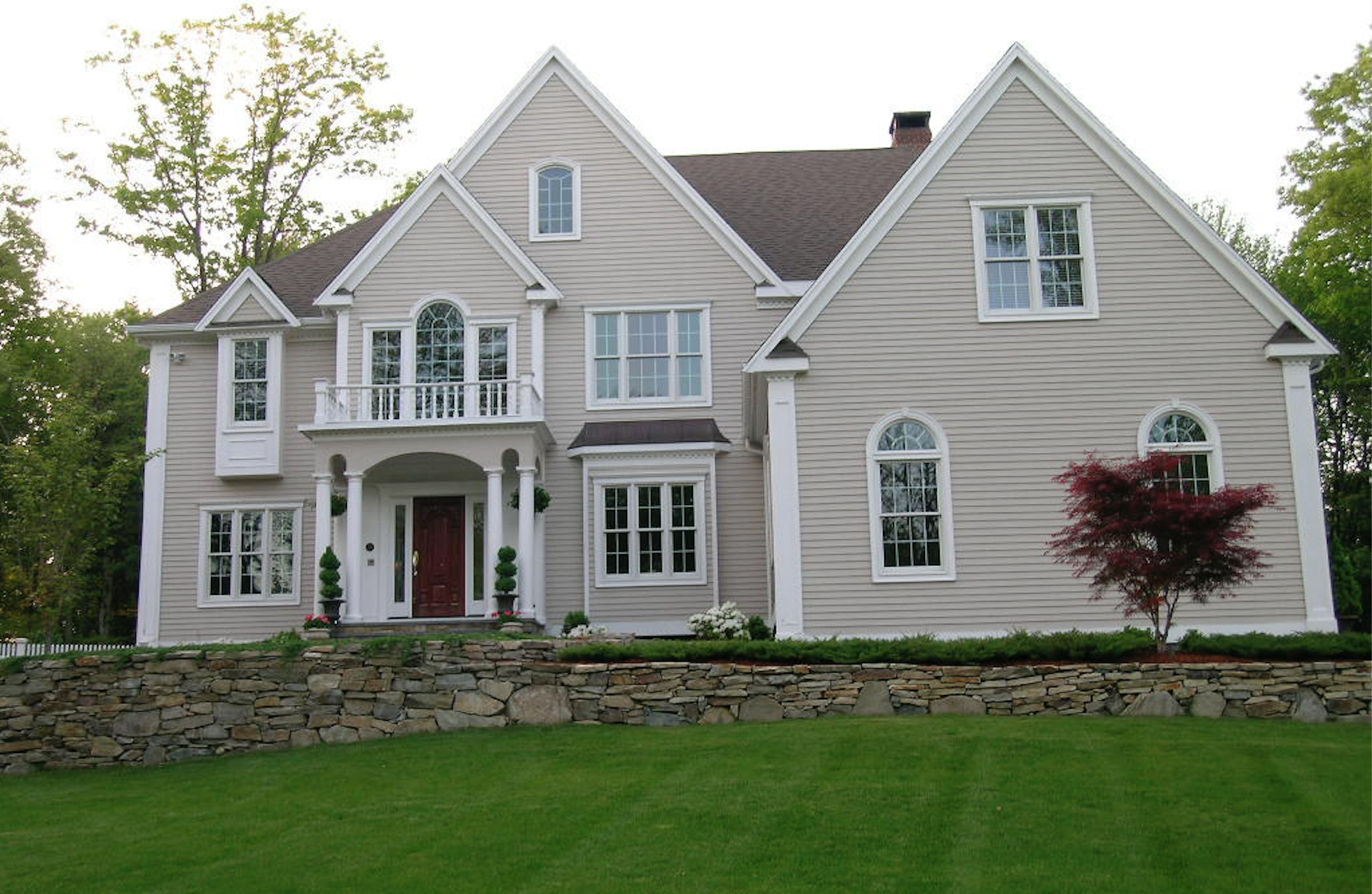 Large two-story house with beige siding, white trim, and a red front door, surrounded by green lawn and trees.