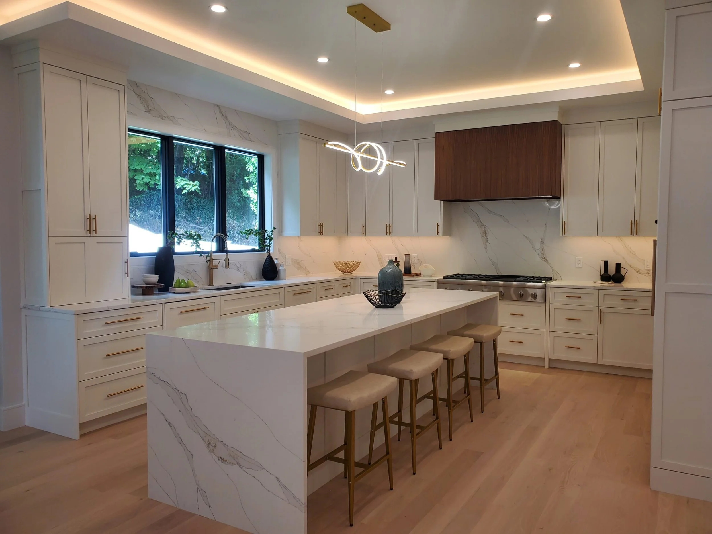 Modern kitchen with white cabinetry, marble countertops and backsplash, a large island with seating, and a window over the sink with trees outside.