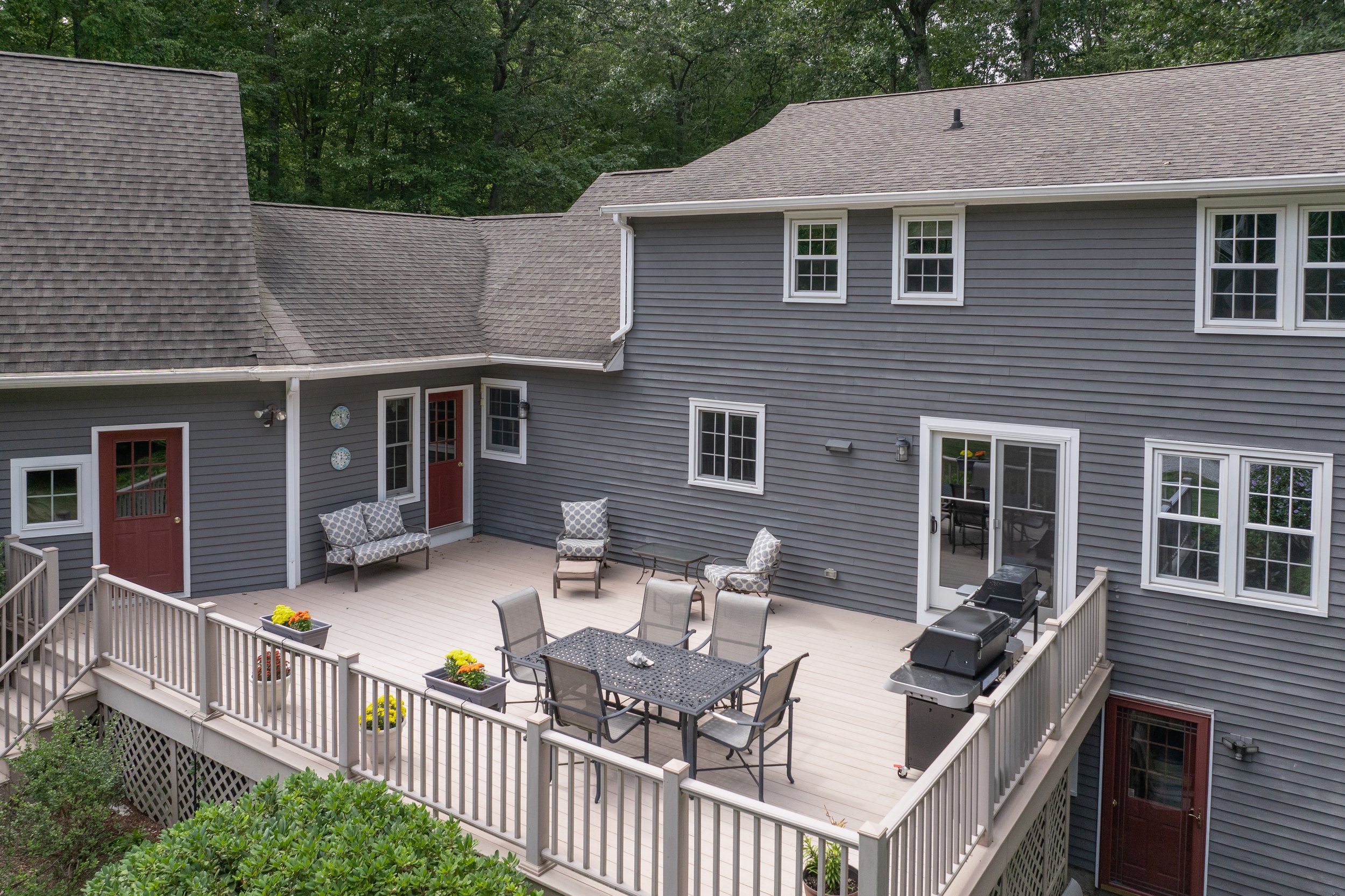 A backyard deck attached to a gray house with sliding glass door, outdoor grill, patio furniture, and surrounded by green trees.