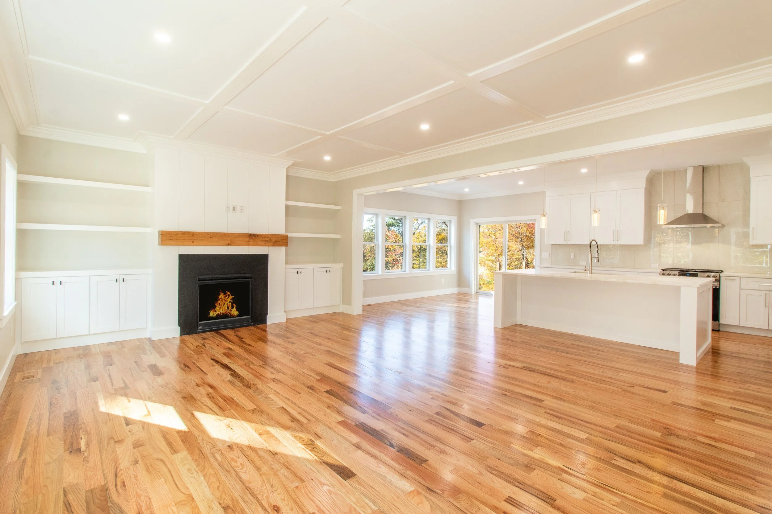 Bright and spacious open-plan living room and kitchen with hardwood floors, white cabinets, and large windows showing autumn trees outside.