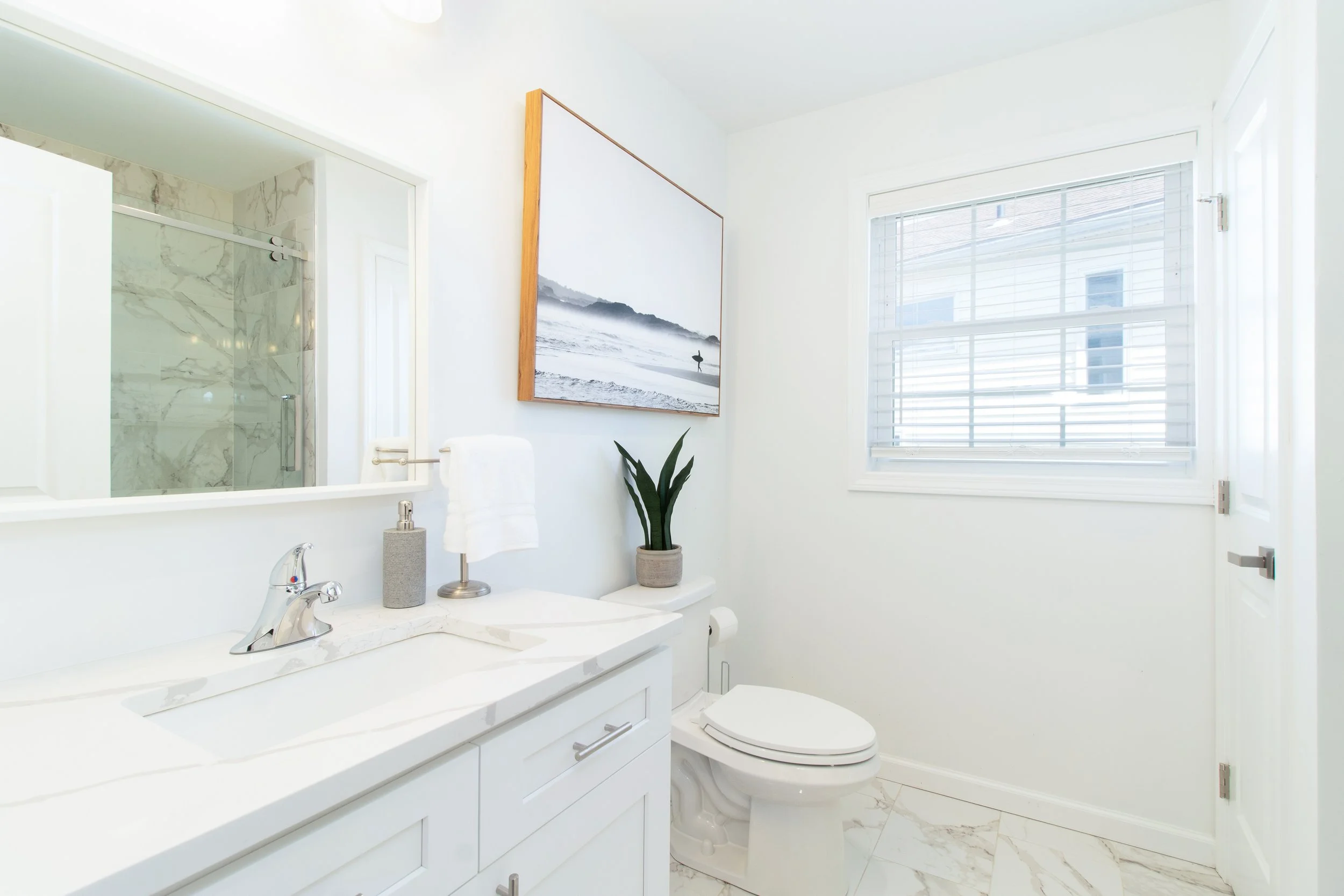 Bright bathroom with white vanity, marble countertop, mirror, potted plant, framed black-and-white landscape photo, toilet, window with blinds, and shower stall with marble walls.