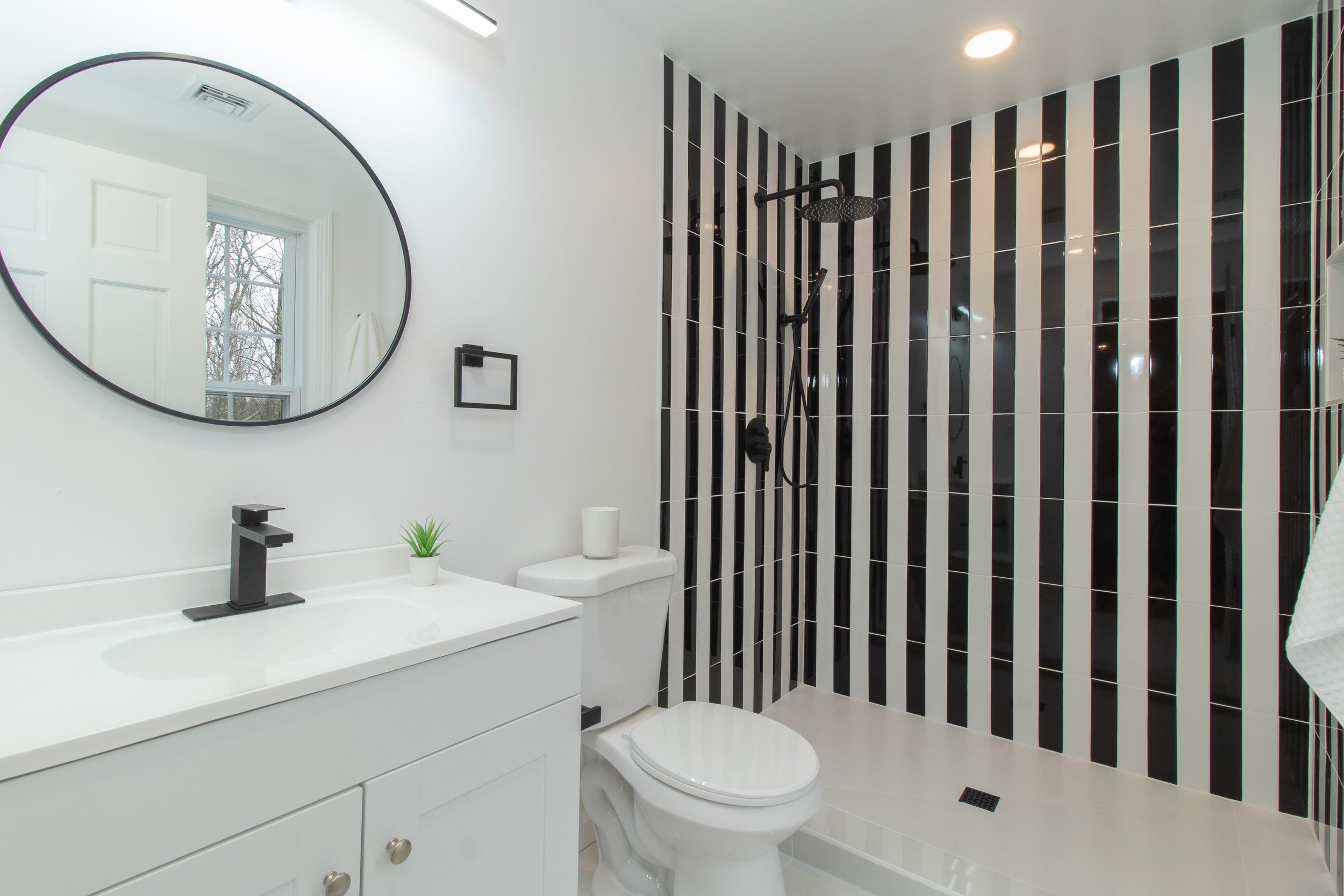 Modern bathroom with black and white striped shower wall, white vanity with black faucet, round mirror, small plant, toilet, and window.