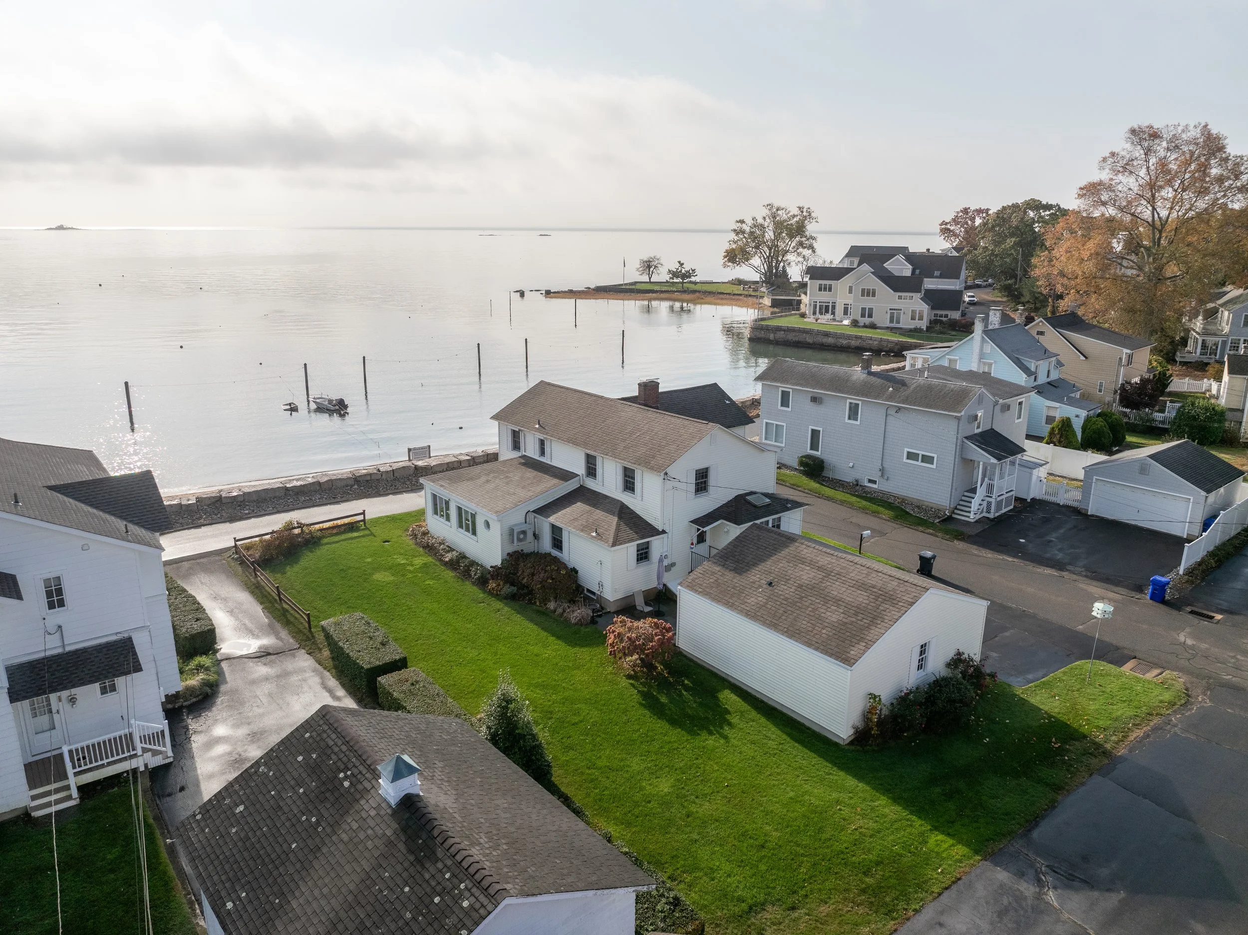 Aerial view of waterfront houses with grassy lawns, boat dock, and calm water with boats and posts at a coastal neighborhood.