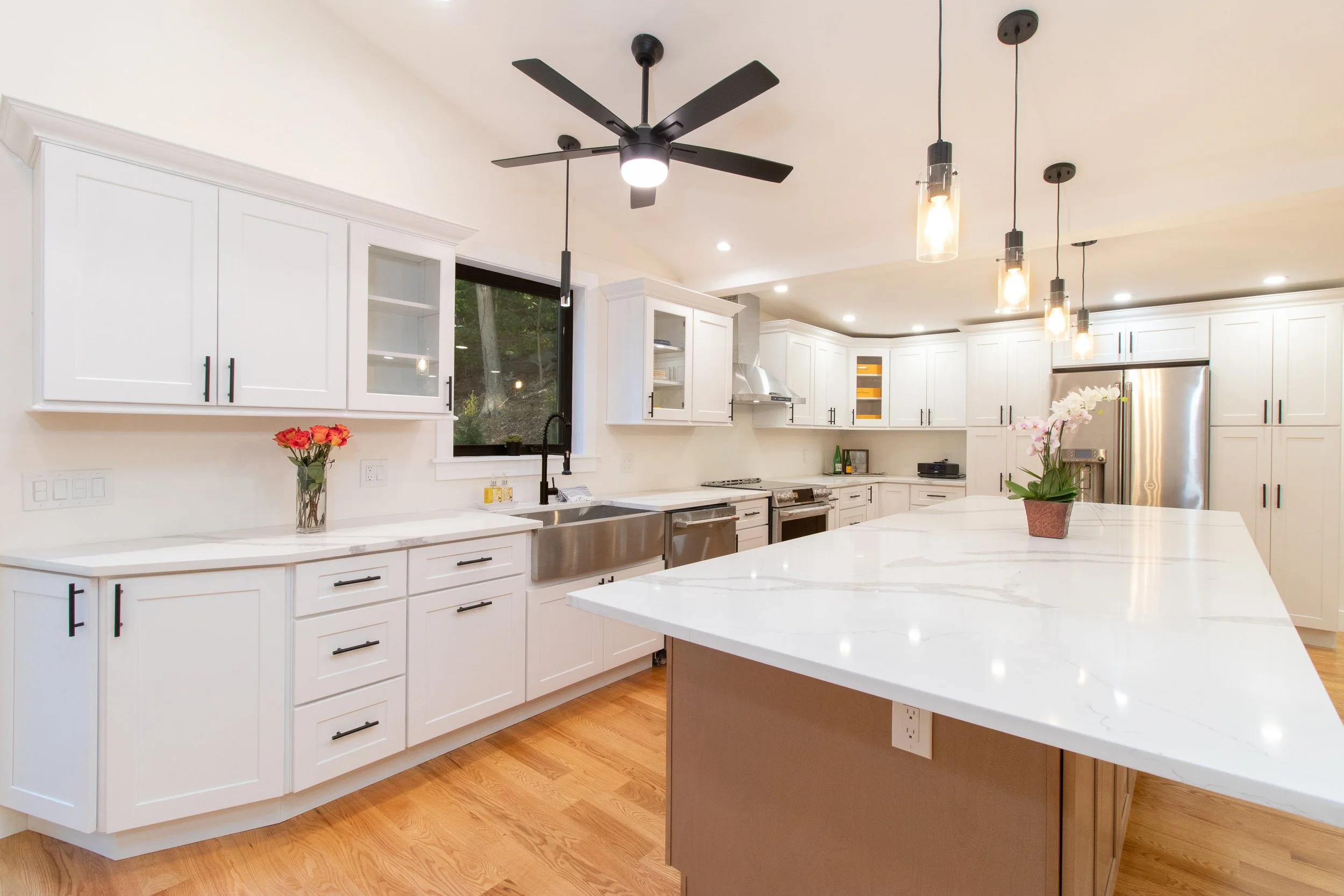Modern kitchen with white cabinets, stainless steel appliances, a large white marble island, black ceiling fan, pendant lights, and flowers