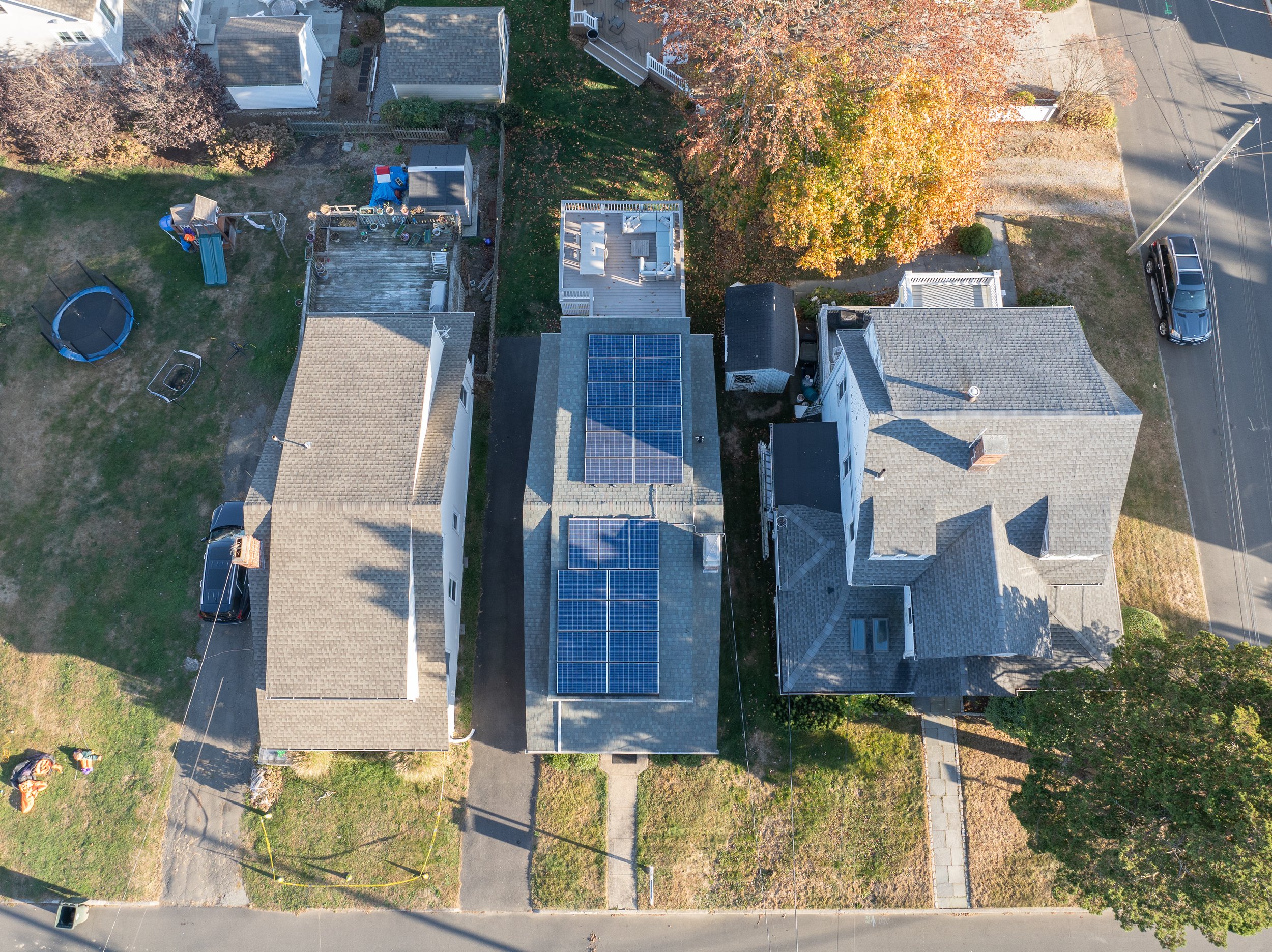 Aerial view of three houses with solar panels on the middle house's roof, surrounded by trees, lawns, and a street with parked cars.