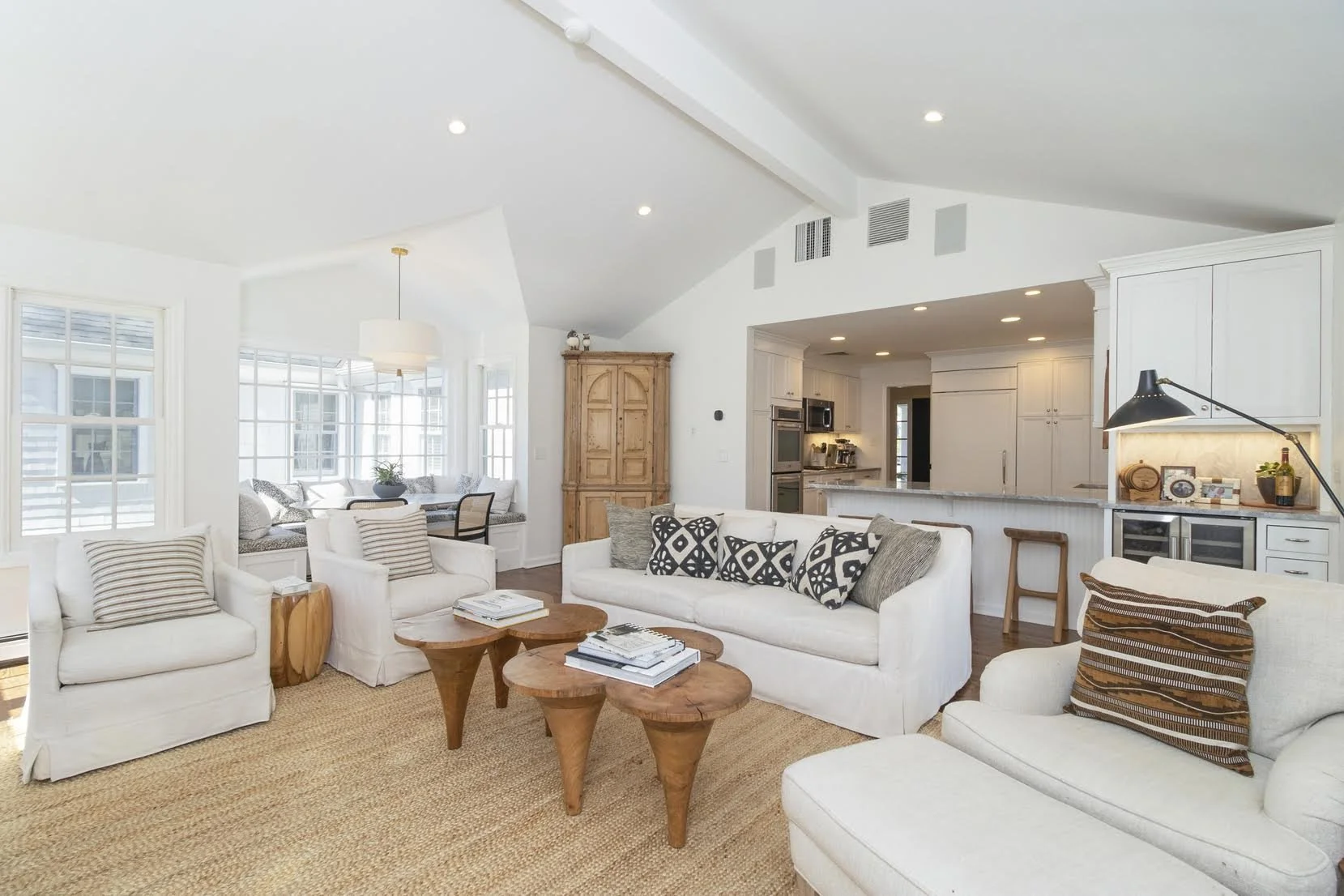 Open-concept living room with white sofas, wooden coffee tables, beige rug, and a kitchen in the background with white cabinets and a breakfast bar.