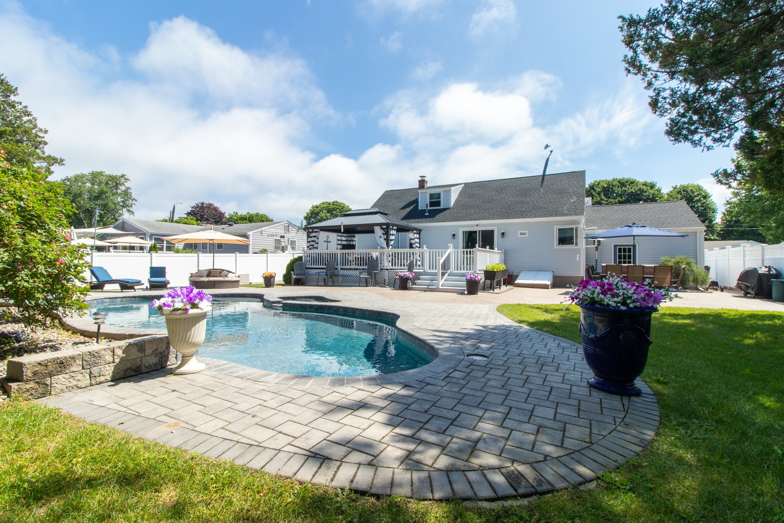 Backyard with swimming pool, patio furniture, and a house under a blue sky with clouds.