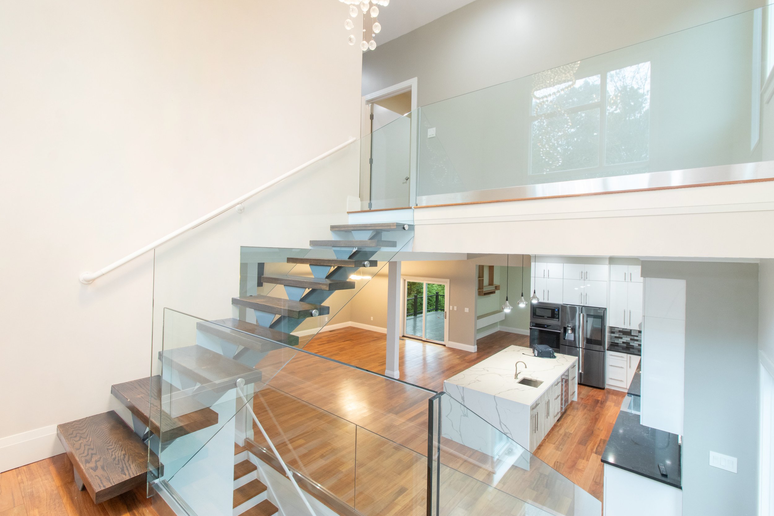 Interior view of a modern two-story house showing a staircase with glass railing leading to a second floor, open kitchen with marble island, and living space with hardwood floors and large windows.