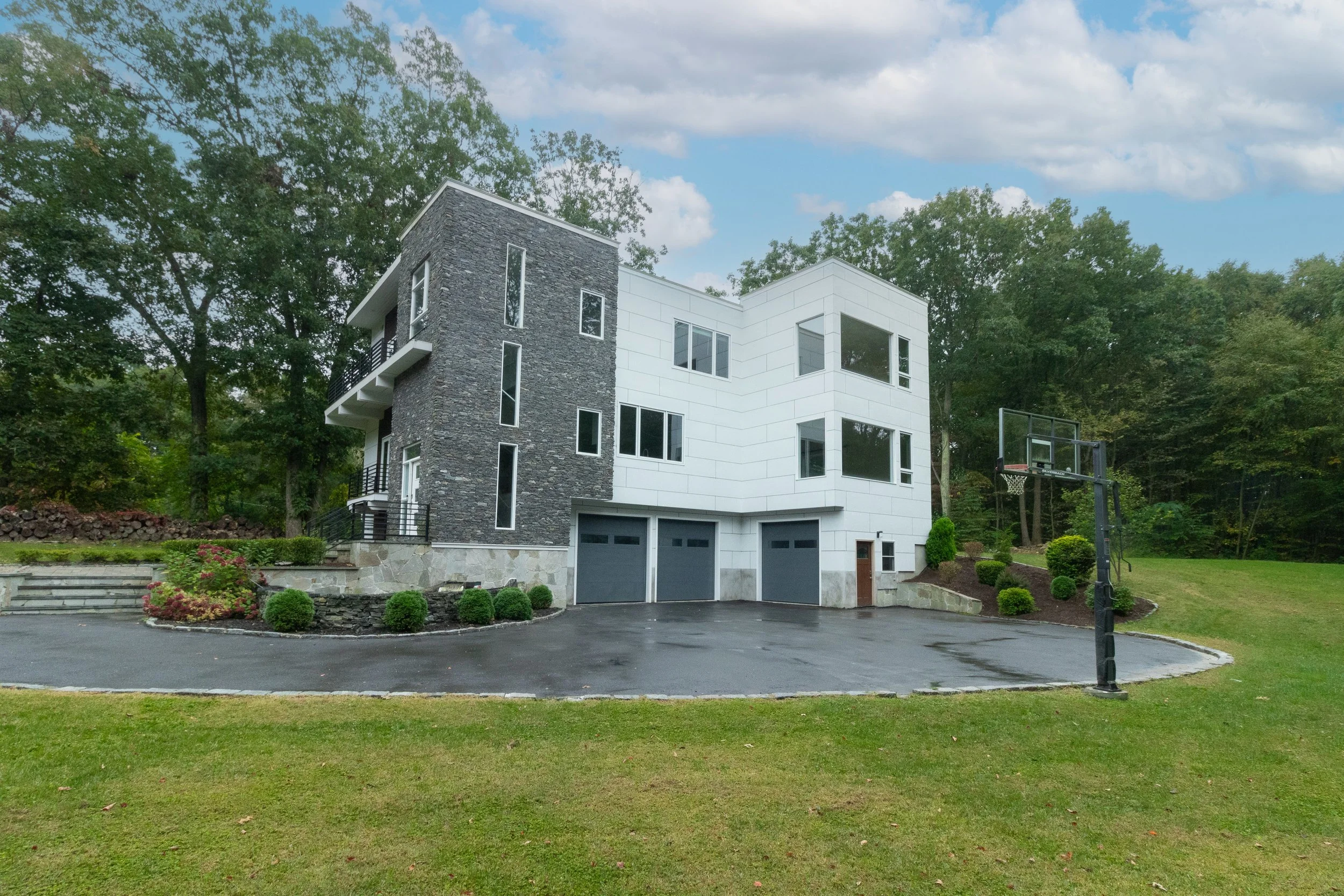 Modern multi-story house with black stone and white exterior, three garage doors, landscaped yard, and a basketball hoop in the driveway, surrounded by green trees and a cloudy sky.