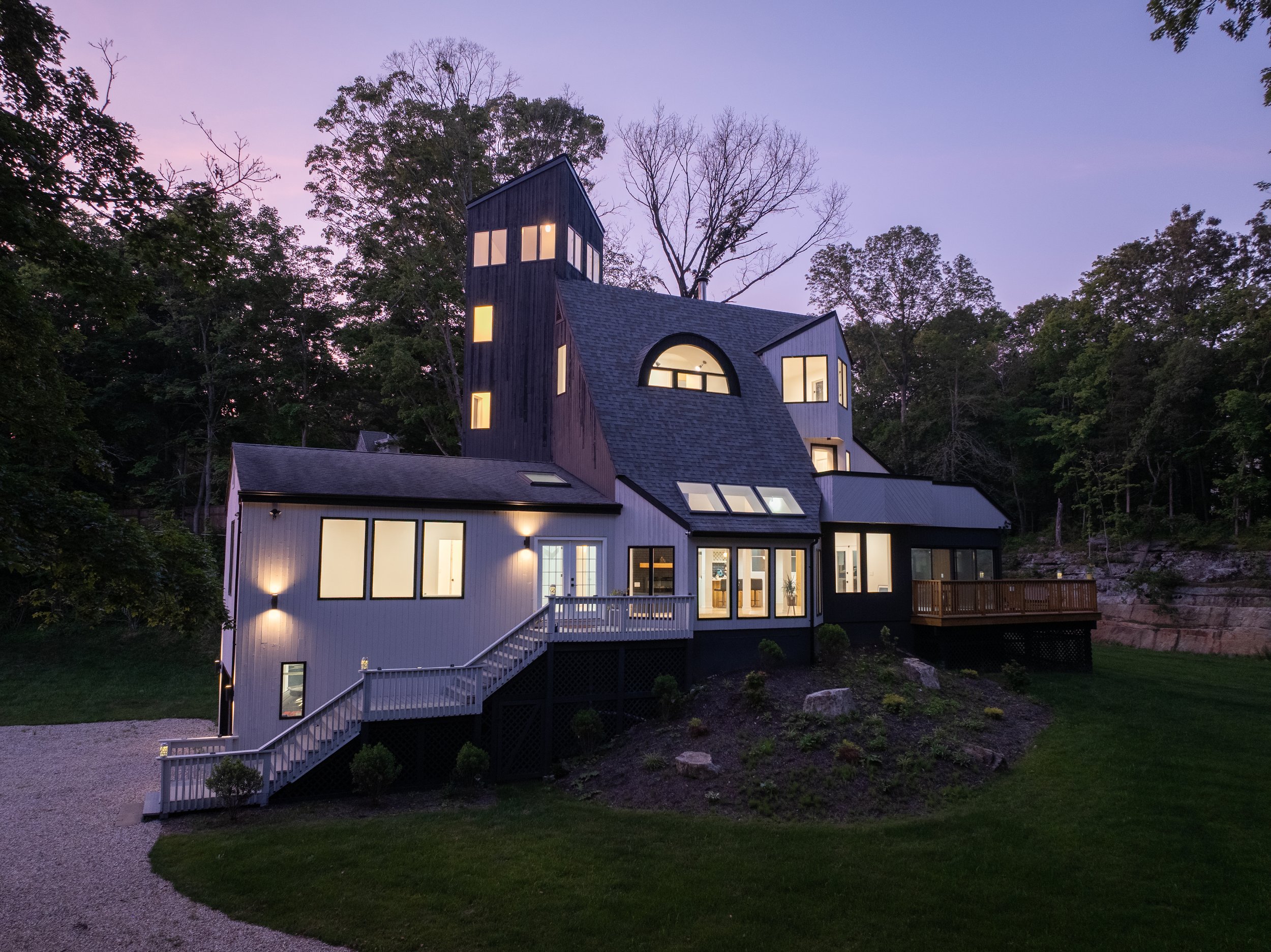 Modern house with multiple levels and angular architecture, illuminated with interior lights, set against a wooded background at dusk in Guilford Connecticut