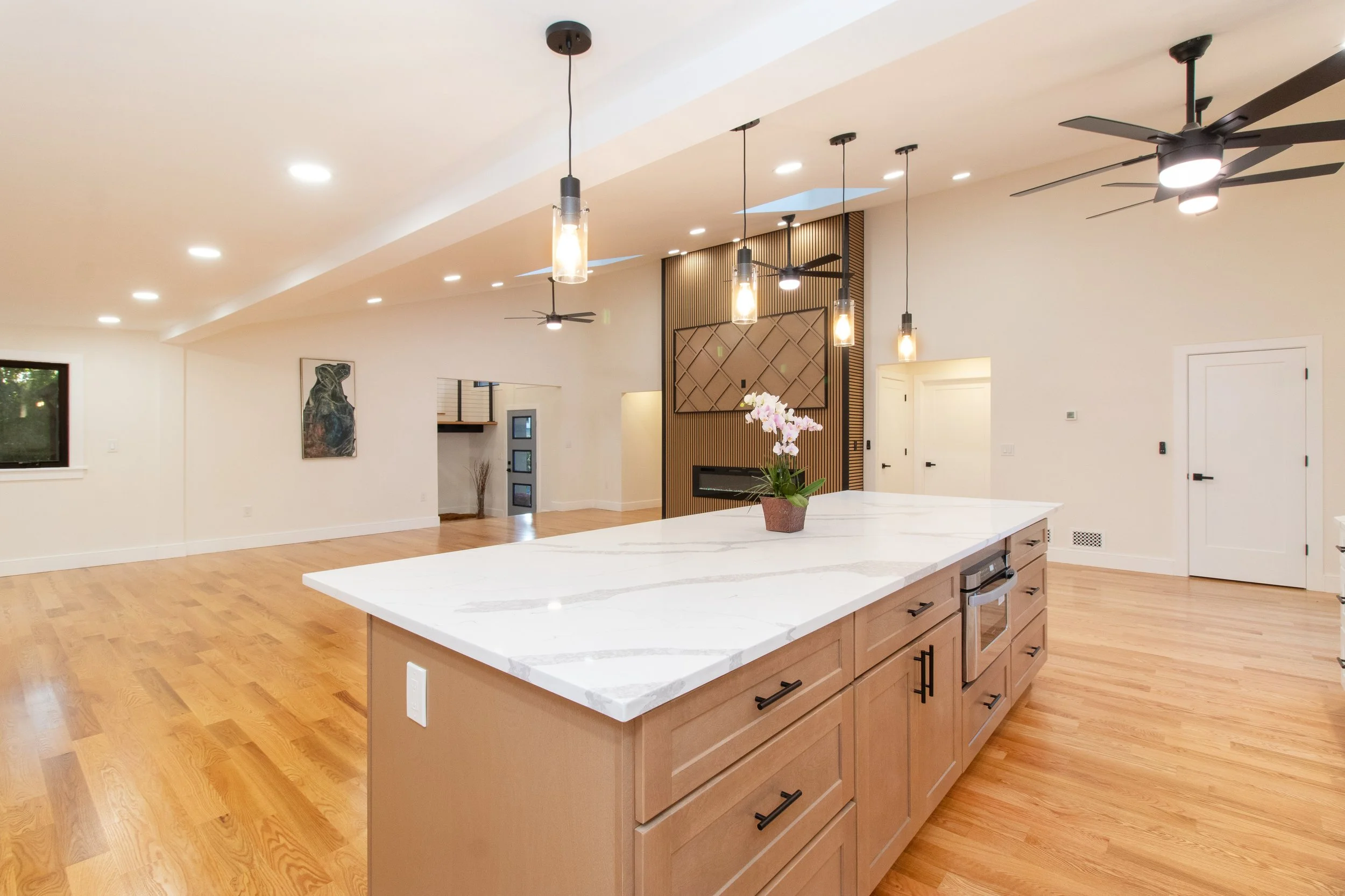 Open-concept kitchen with a large white marble island, wooden cabinetry, hardwood flooring, modern pendant lighting, ceiling fans, and a decorative feature wall with a built-in fireplace.