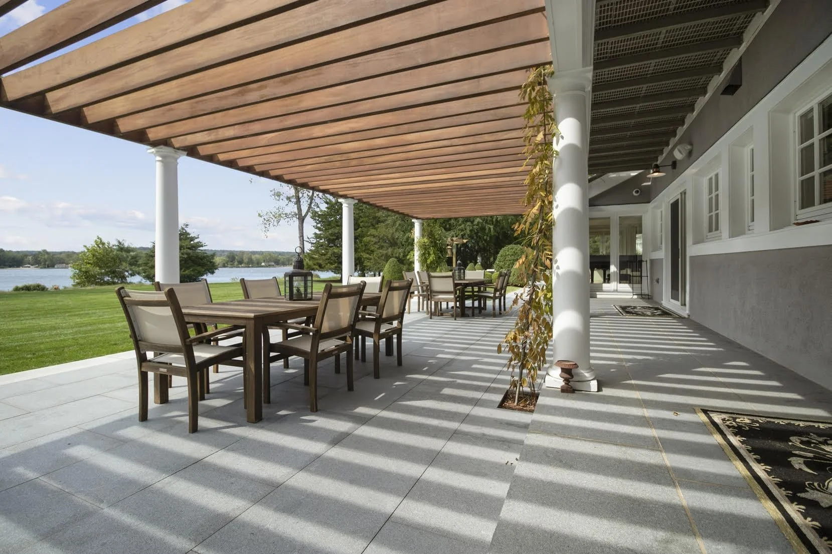Large outdoor patio with wooden dining tables and chairs, shaded by a wooden pergola, overlooking a lake with green grass and trees in the background.