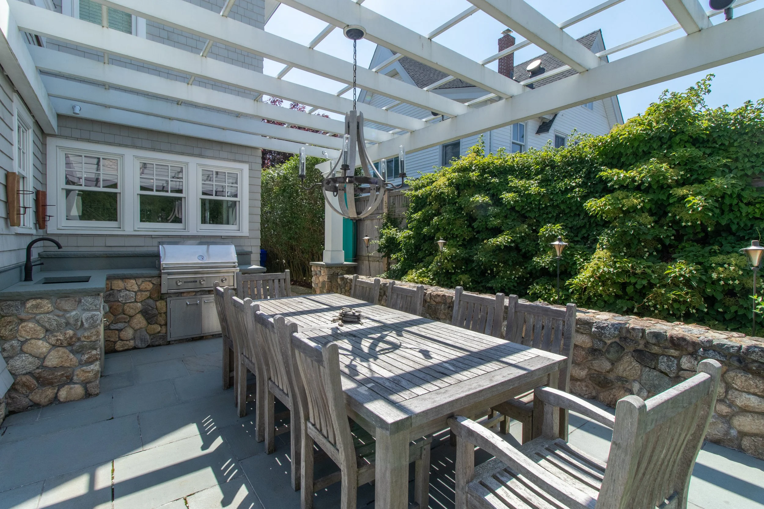 Outdoor patio area with wooden dining table and chairs, built-in outdoor kitchen with grill, surrounded by lush greenery, and a chandelier hanging from an open pergola structure