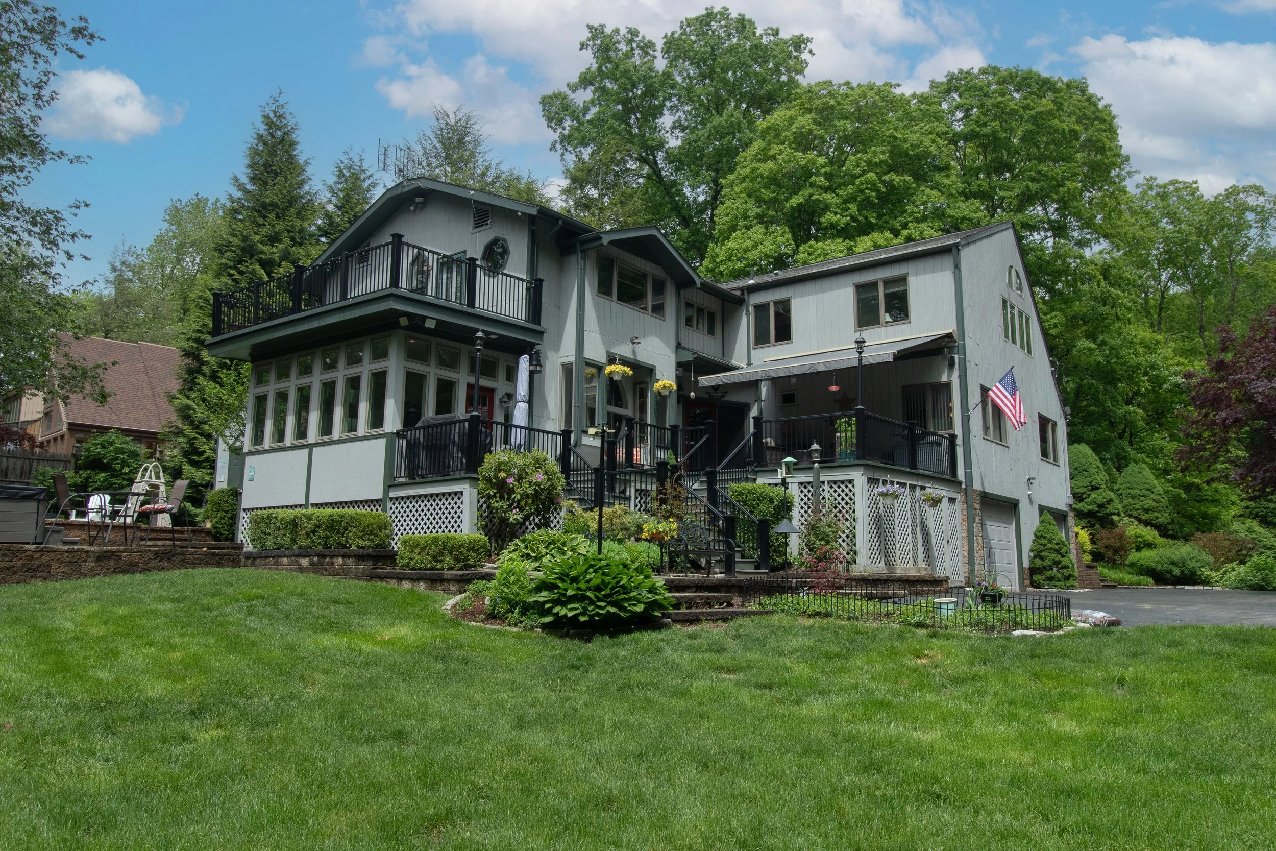 A large multi-story house painted gray with black railings, surrounded by a well-maintained lawn and lush green trees.
