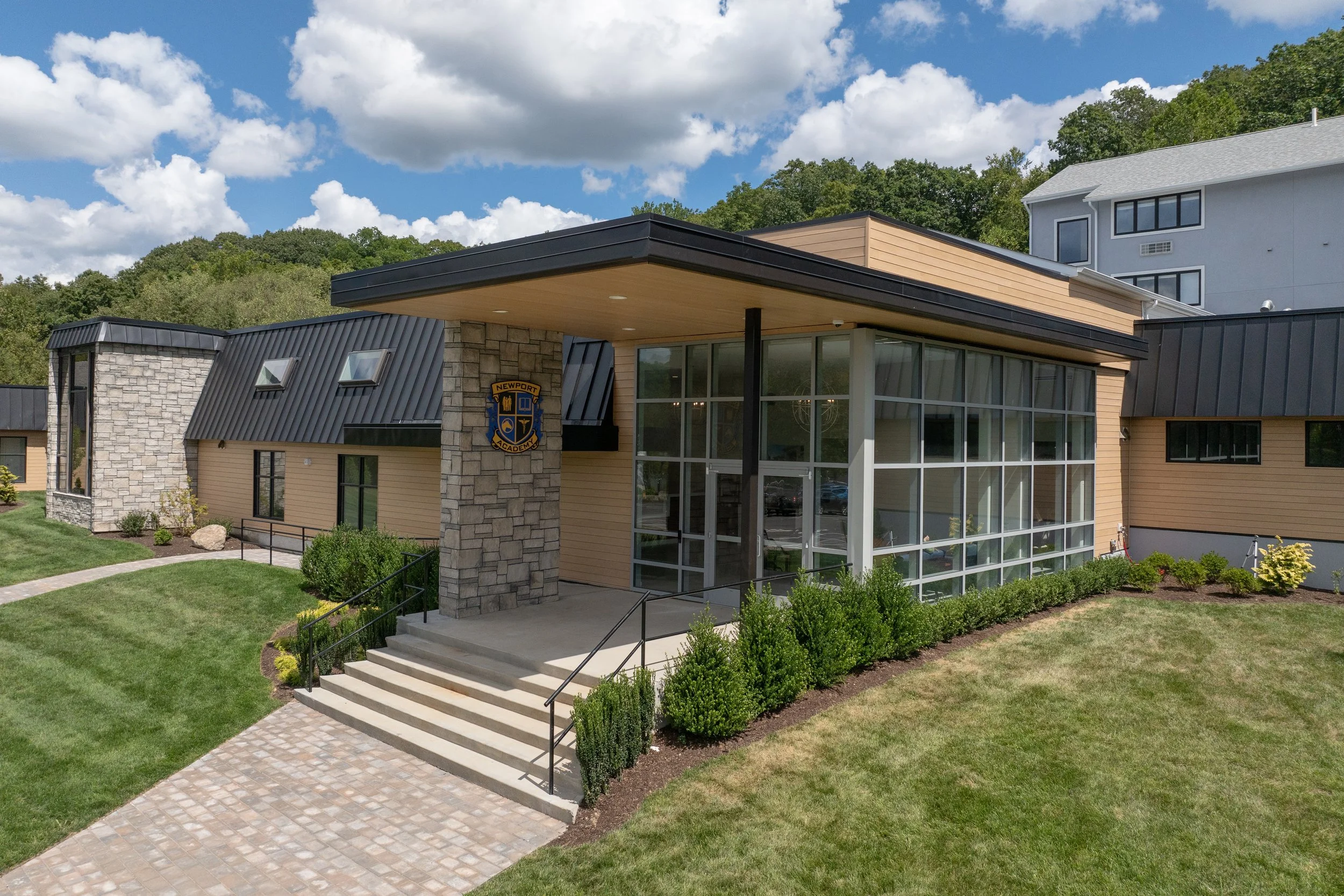 A modern school building with beige siding, large glass windows, and a stone and metal accent wall displaying a crest that reads "Newport Academy." The entrance has a small set of concrete steps, lined with black metal railings, leading to a covered porch area. Well-maintained green lawn and shrubs surround the building, with a paved walkway leading to the stairs.