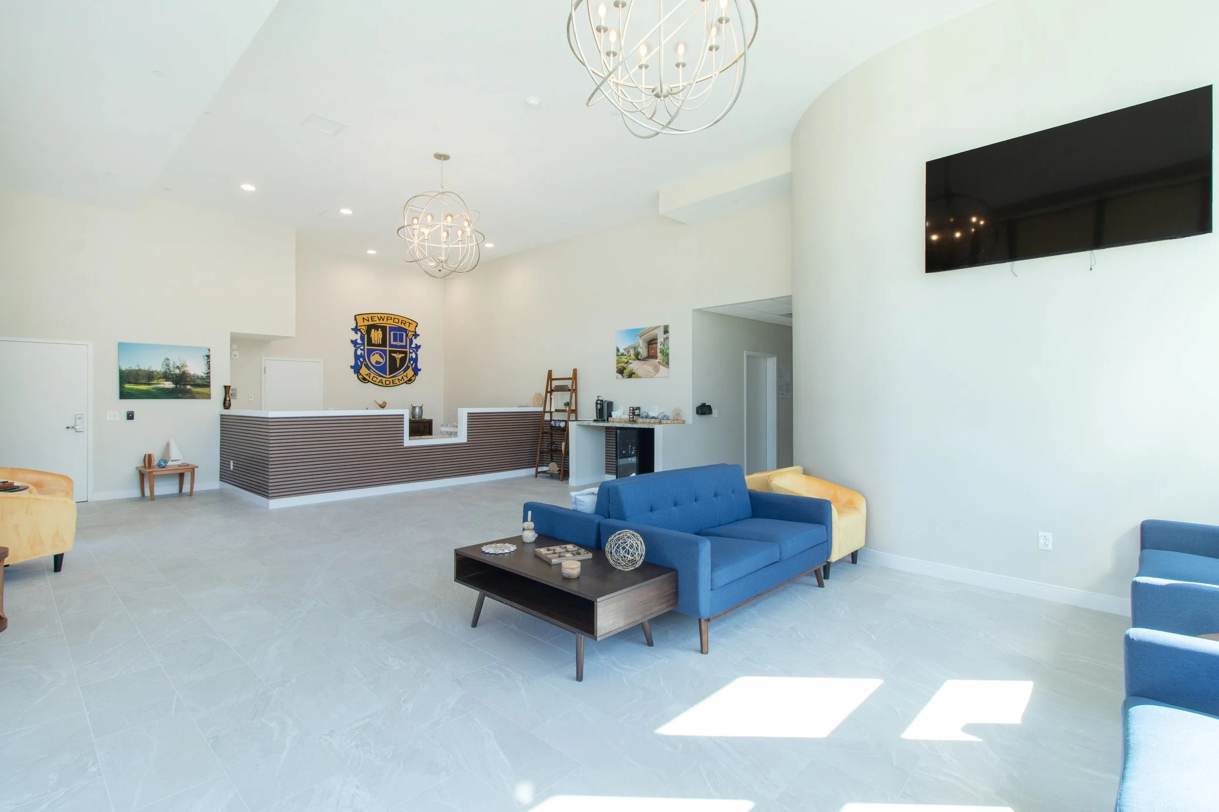 Modern hotel lobby with a reception desk, multiple blue and yellow couches, a wall-mounted TV, chandeliers, and artwork on white walls.