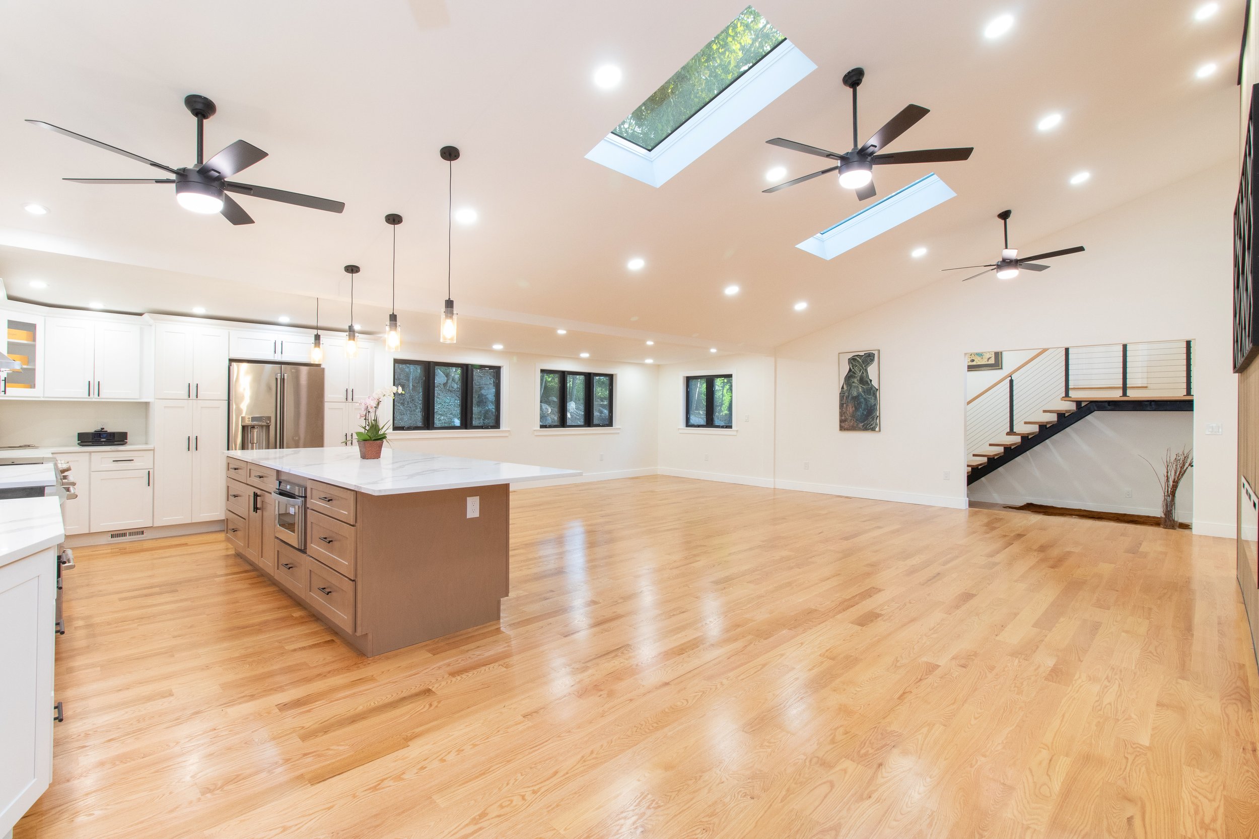 Open and spacious living room and kitchen area with hardwood floors, white cabinets, and large windows. Kitchen has an island with pendant lights above, and ceiling fans and skylights are visible on the ceiling.