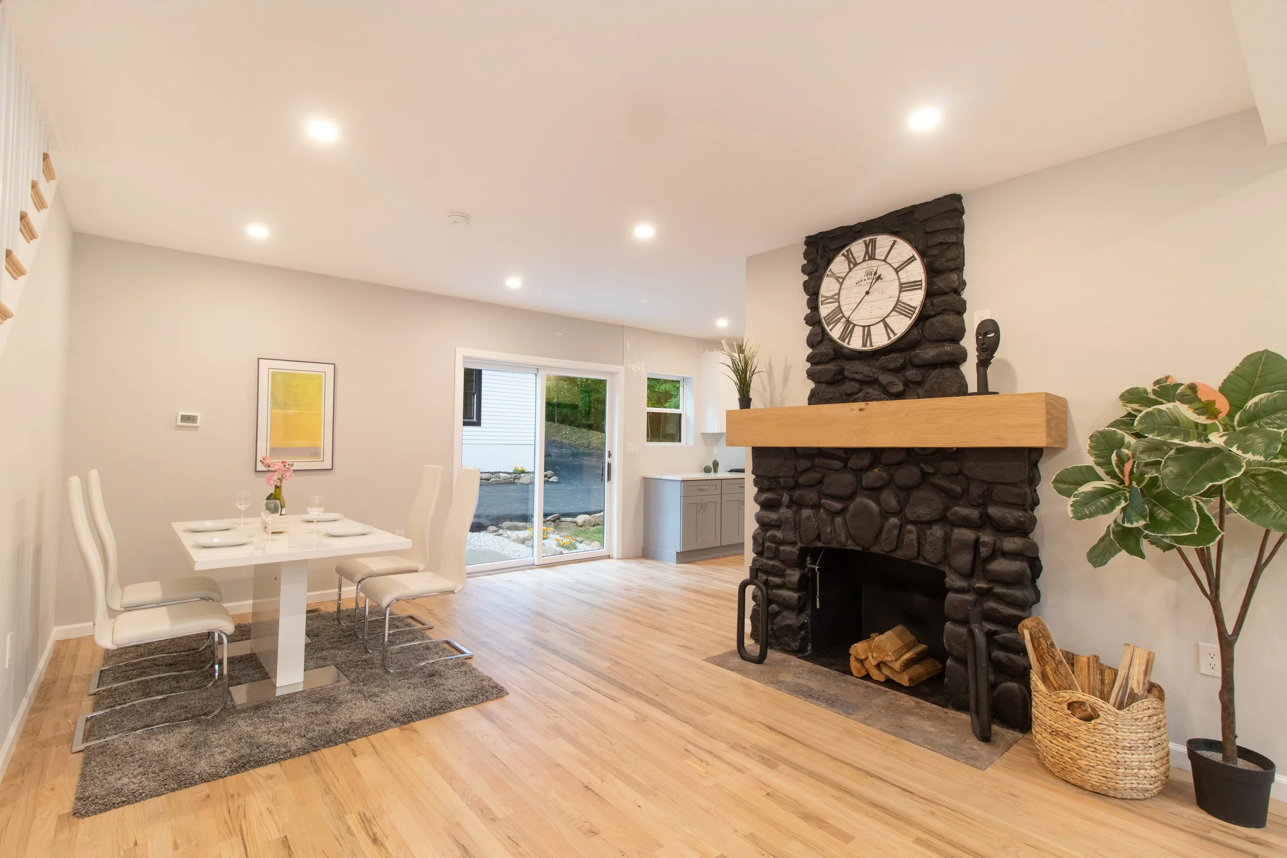 Modern living room with a stone fireplace, wooden mantel, large clock, potted plant, dining table with chairs, sliding glass door, and hardwood floors. Real Estate Photography in Guilford Connecticut