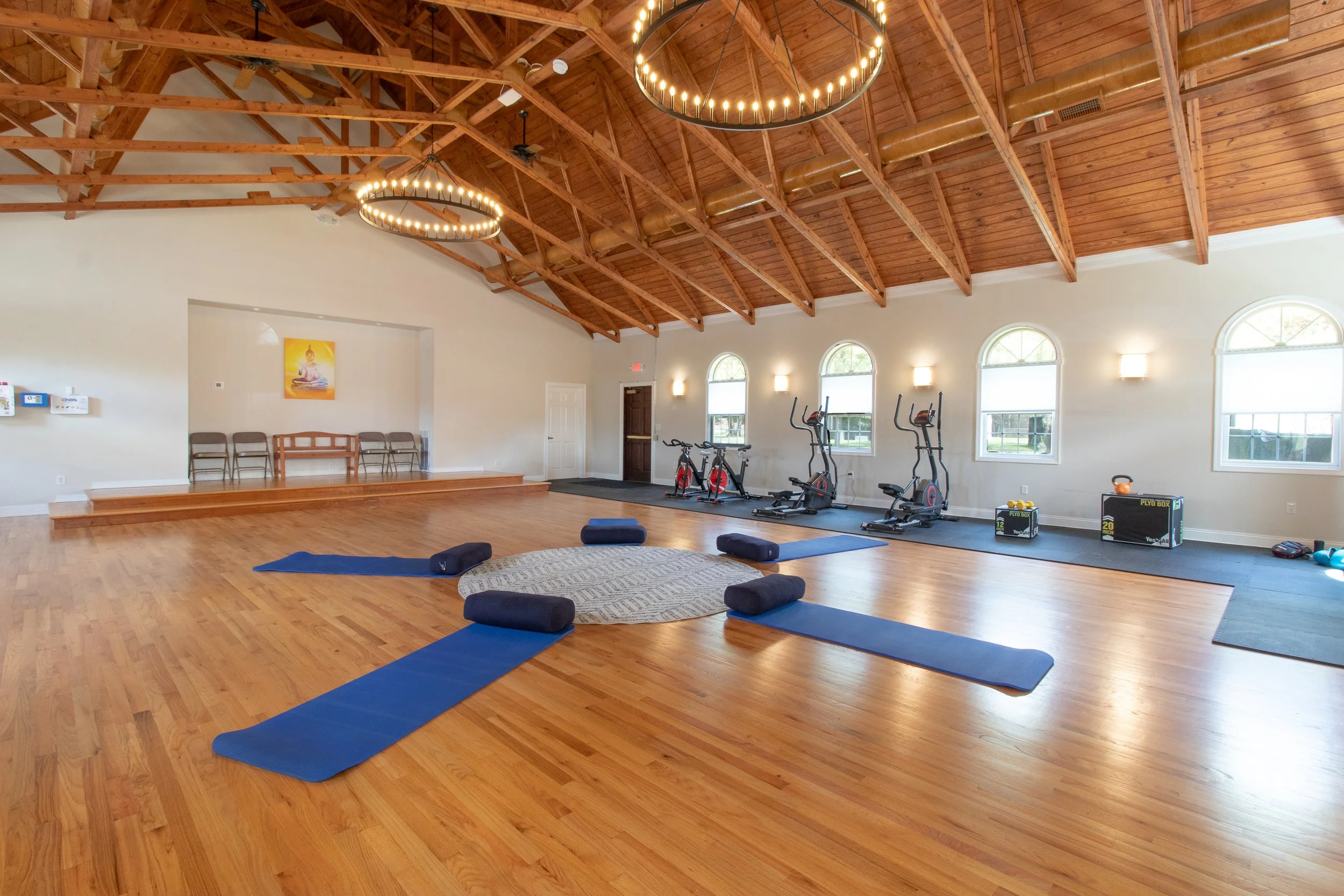 A spacious indoor wellness or yoga studio with wooden floors and a high wooden ceiling. There are fitness and exercise equipment on the right side, including stationary bikes, ellipticals, and a small workout area with weights. In the foreground, there are yoga mats, bolsters, and a circular rug, suggesting a yoga or meditation space. The room is well-lit with natural light coming through arched windows and has modern lighting fixtures on the walls.