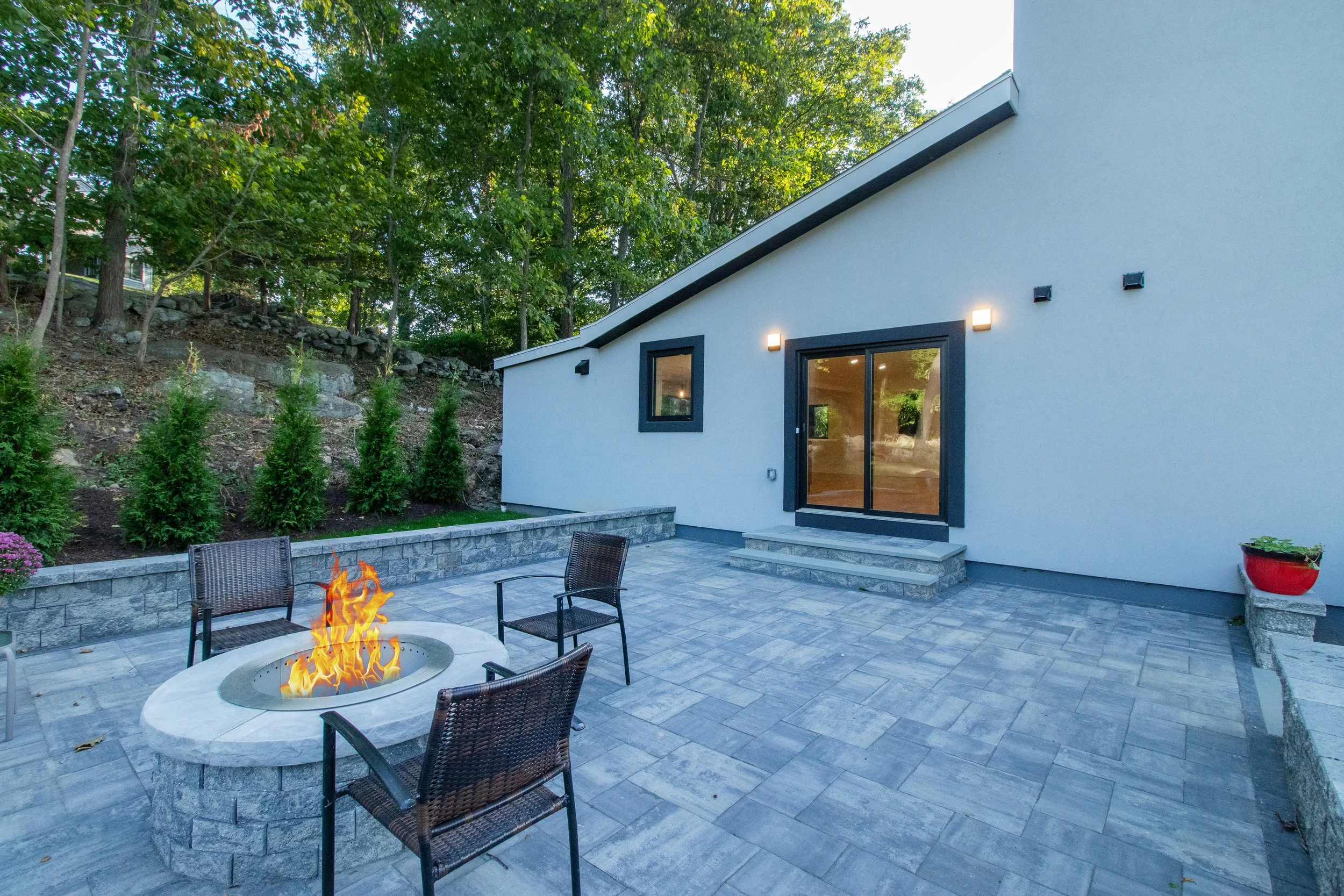 Modern backyard patio with a fire pit surrounded by four chairs, overlooking a house with sliding glass doors, outdoor lighting, and a stone retaining wall with plants and trees in the background.