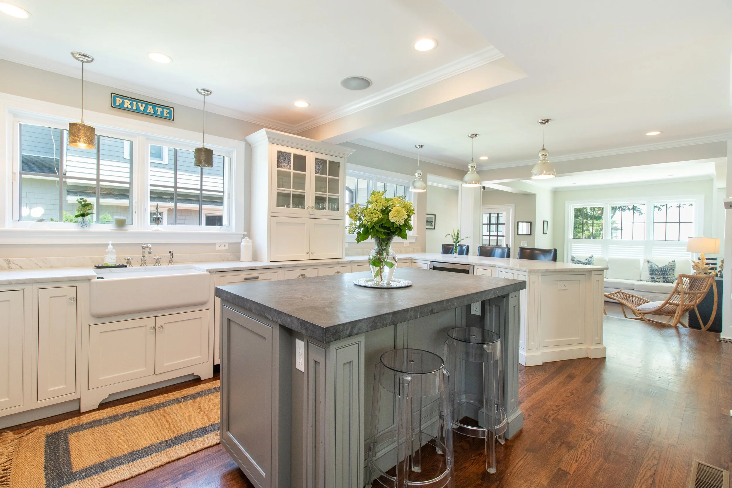 Bright kitchen with white cabinets, gray island, and wood flooring. A vase of flowers is on the island. There are windows, pendant lights, and a cozy seating area in the background.