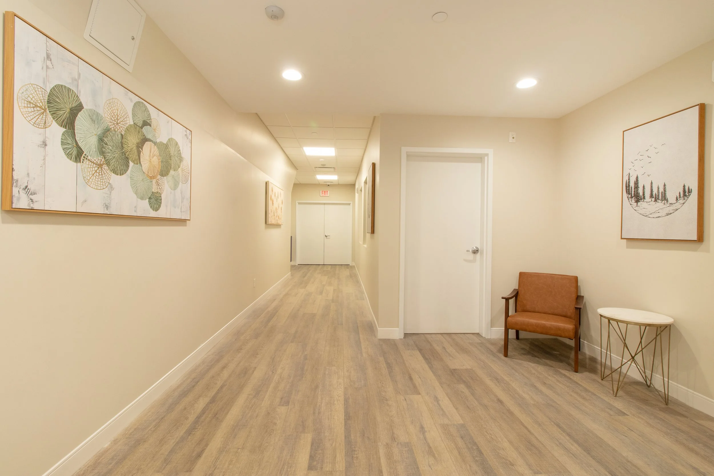 Empty hallway with beige walls, wooden flooring, and wall art, featuring a brown armchair and a small side table near the wall. Real Estate Photography in Seymour Connecticut