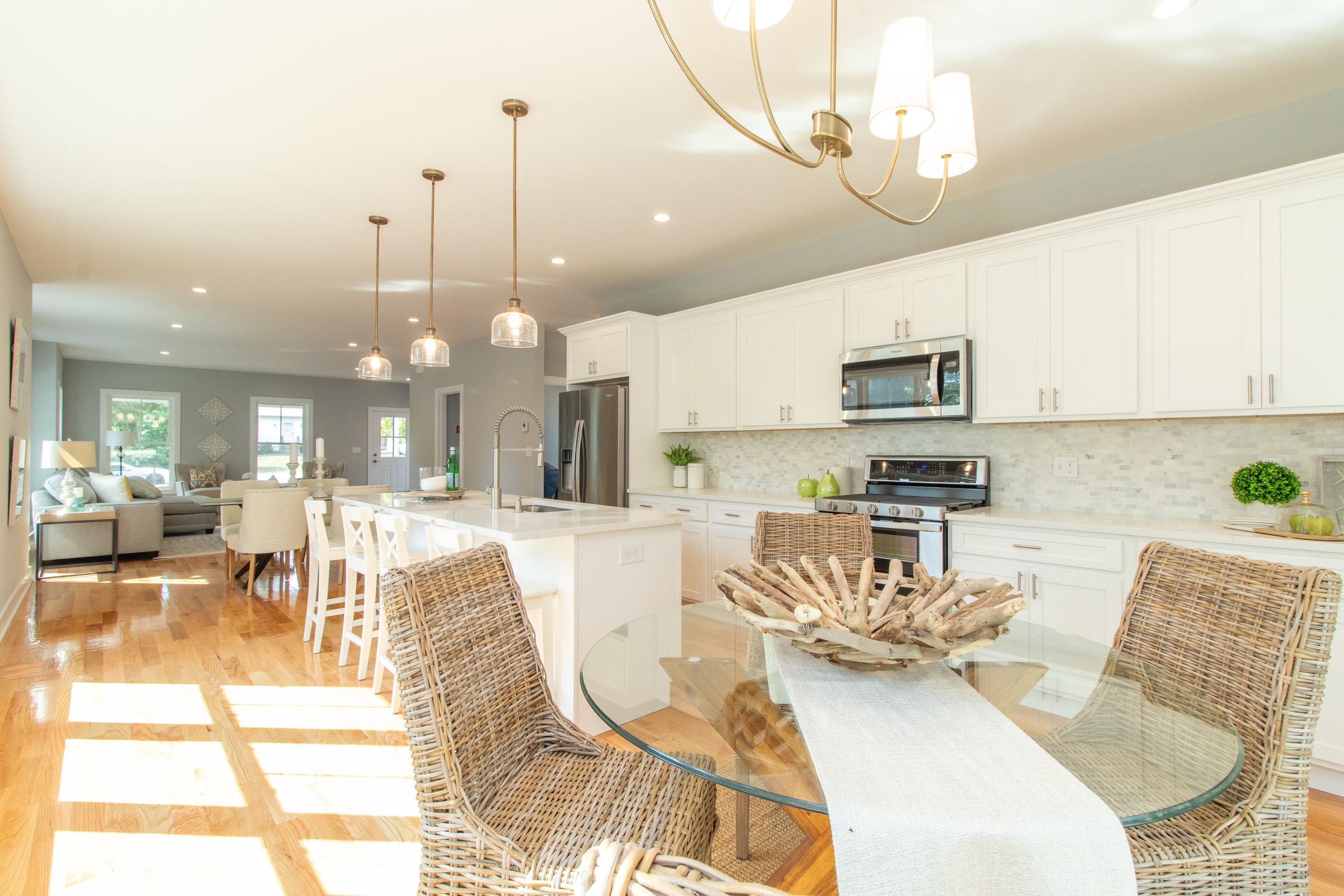 Open-concept kitchen with white cabinets, stainless steel appliances, a glass-top dining table with wicker chairs, a kitchen island with white bar stools, and a living room with sofas and large windows.