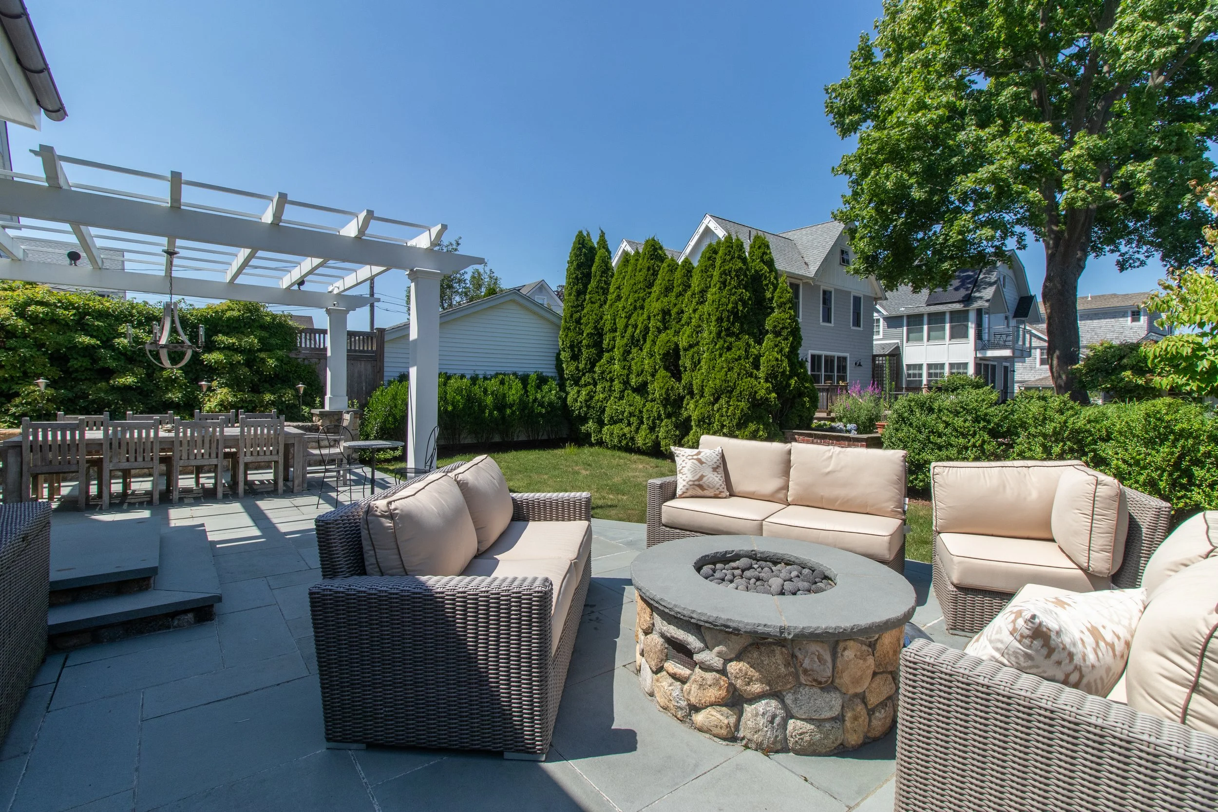 Backyard patio with outdoor furniture, a fire pit, a pergola with a chandelier, lush green trees and shrubs, and houses in the background under a clear blue sky.
