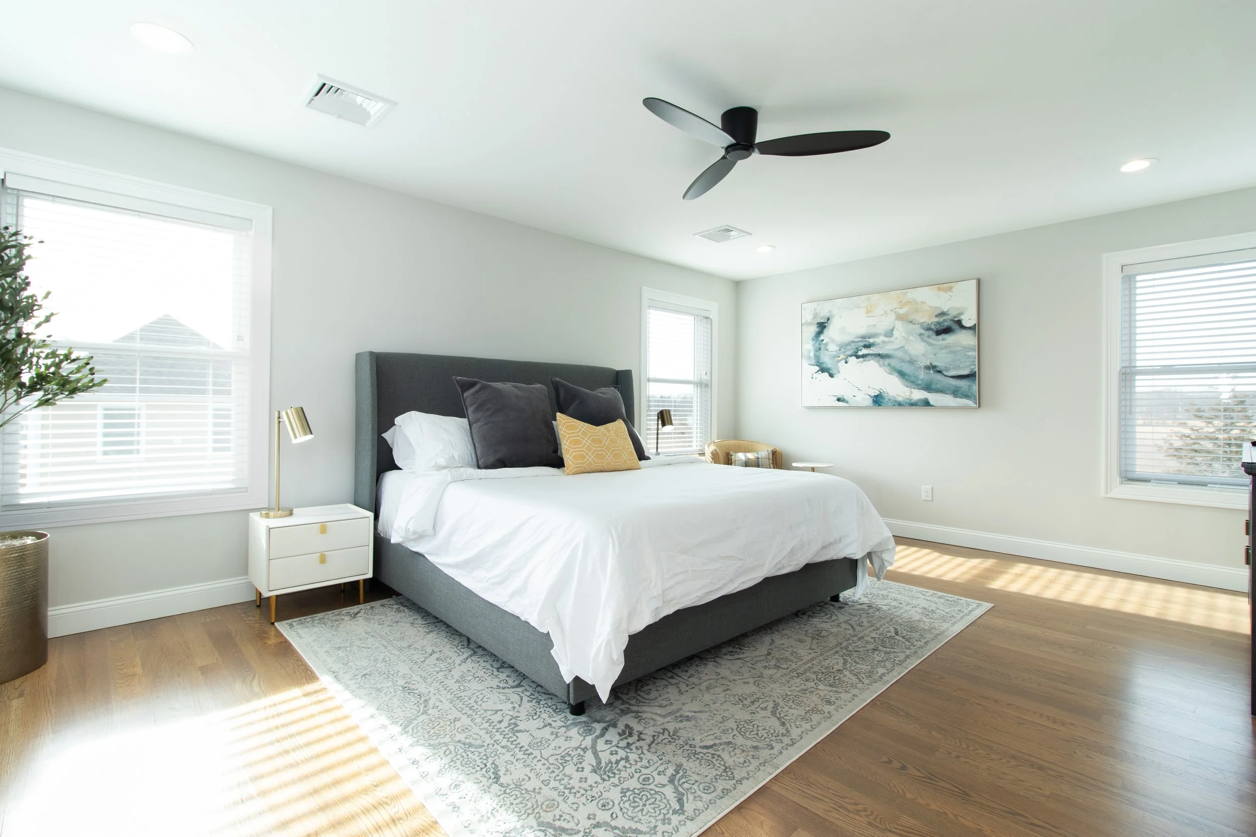 Bright bedroom with large bed, gray upholstery, white bedding, and dark gray pillows. Two gold-accented bedside lamps, a white nightstand, a patterned rug, and modern artwork on the wall. Multiple windows with blinds and a ceiling fan.